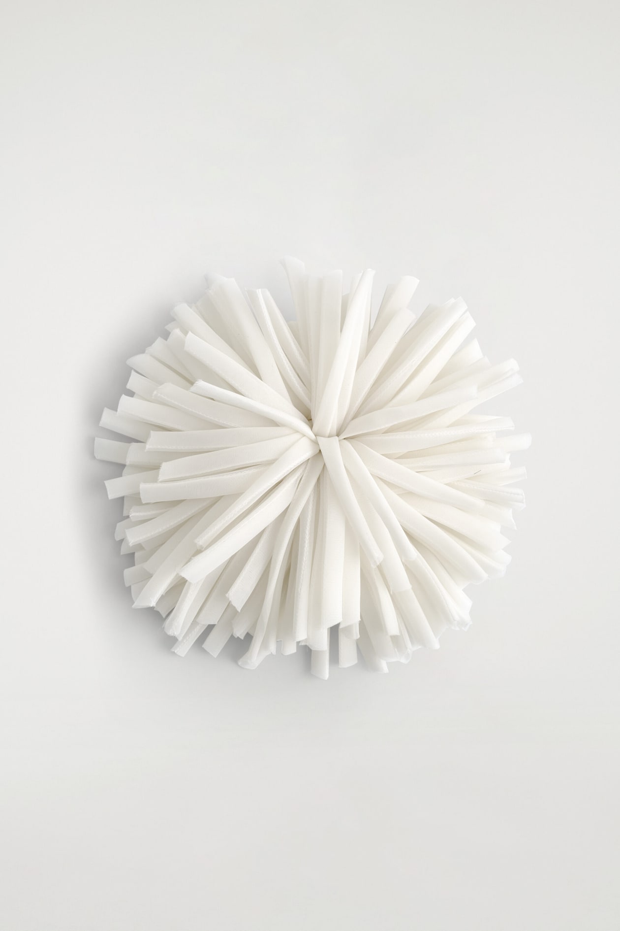 A white floral rosette brooch is shown from the front, featuring numerous thin, semi-sheer fabric ribbons radiating from a central point to form a textured, spherical pom-pom shape.