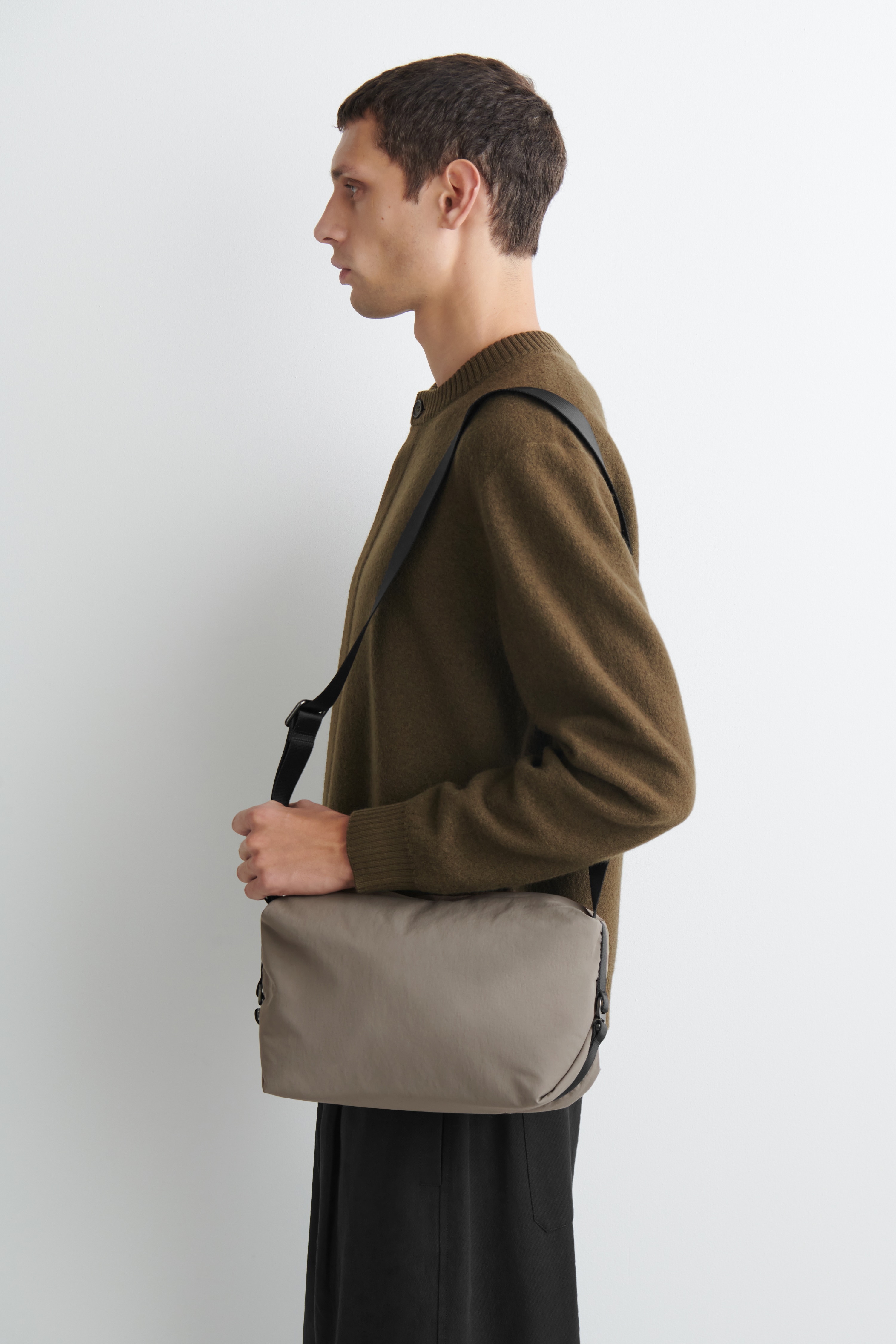 A taupe nylon crossbody bag is shown from the front, featuring a soft rectangular shape and a long, adjustable black webbing strap extending upwards. The bag is finished with small black buckles and hardware securing the straps on each side.