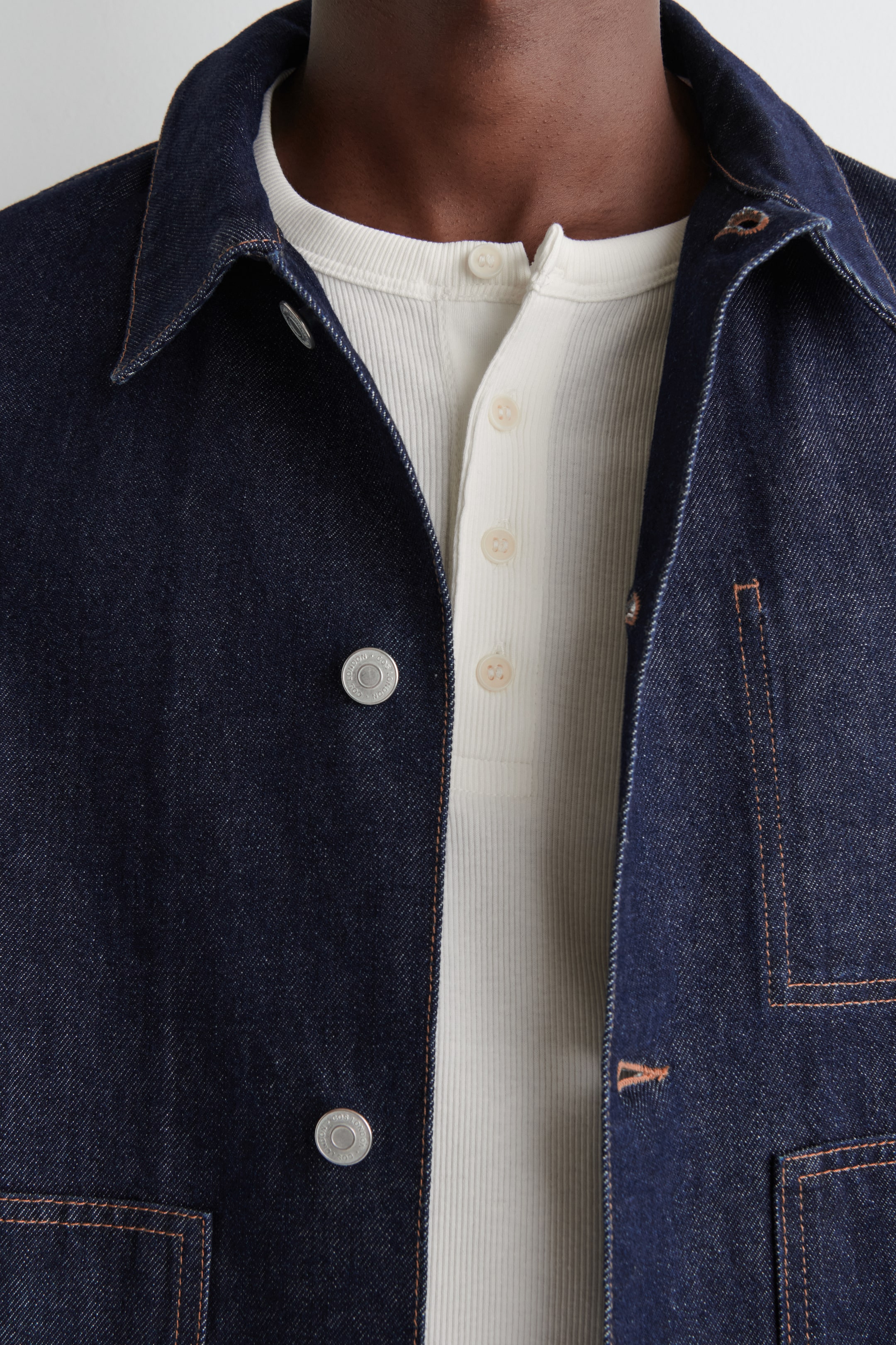 View larger image: A dark denim jacket featuring silver-toned snaps and contrast stitching is being worn open over a light-colored, ribbed Henley shirt.