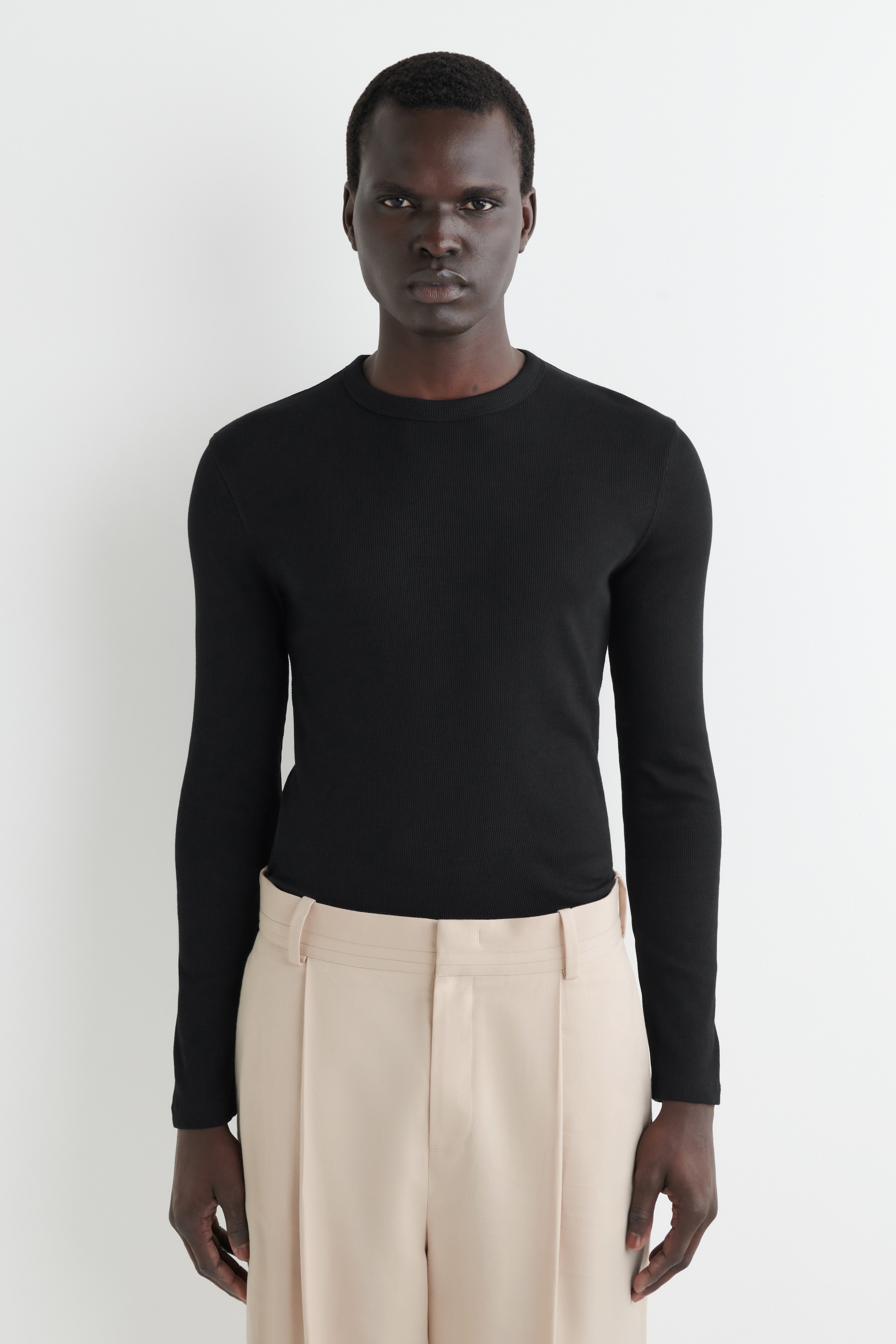 Ver imagen más grande: Un top negro de manga larga acanalado se presenta de frente, con un cuello redondo y un ajuste entallado. Se lleva metido dentro de unos pantalones beige claro, mostrando su longitud y forma.