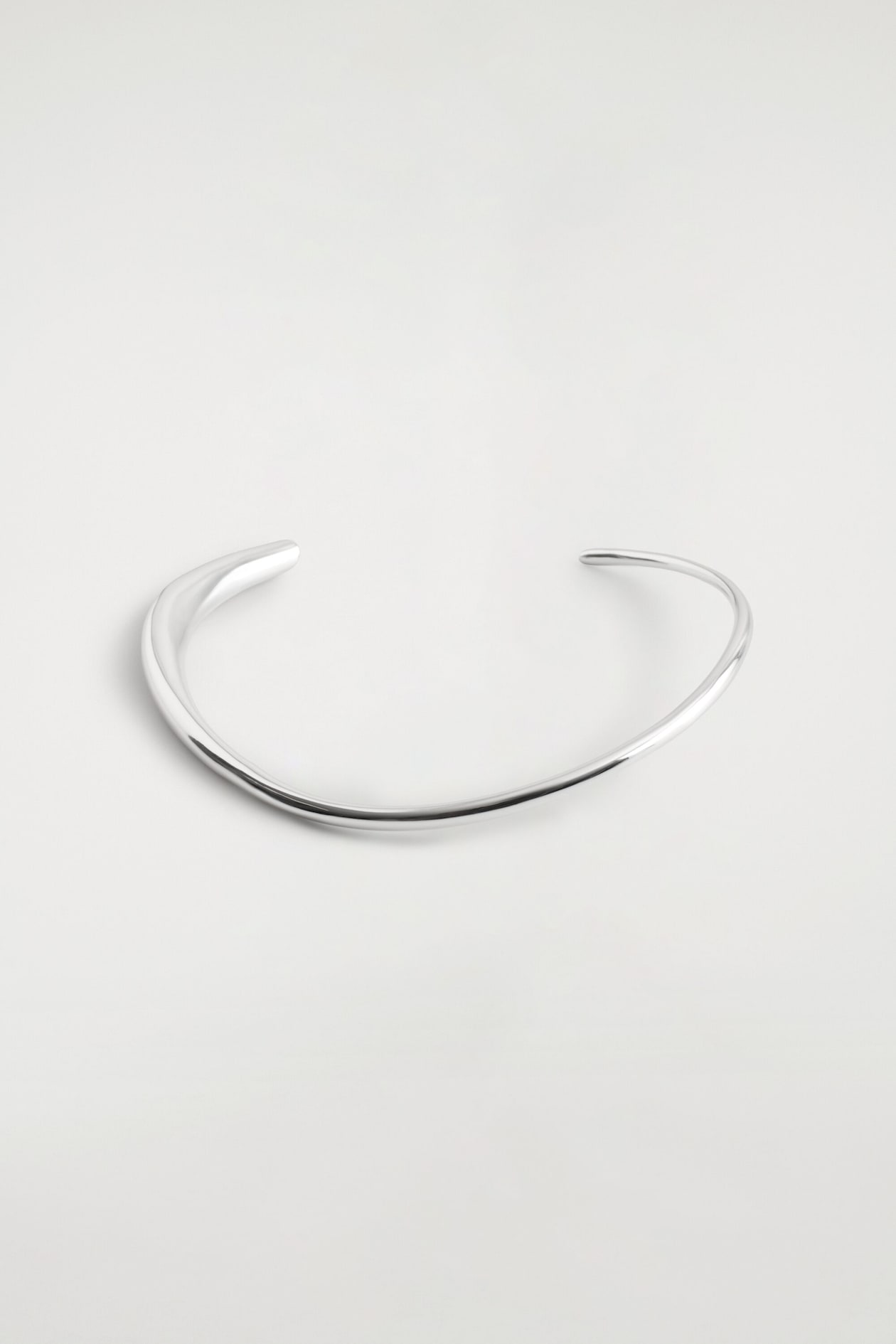 Un collier à col ouvert de teinte argentée repose à plat, mettant en valeur son design minimaliste et sculptural. La bande en métal poli présente une courbe asymétrique et organique qui s’effile en une fine extrémité arrondie d’un côté et s’élargit en une forme plus épaisse et aplatie de l’autre.