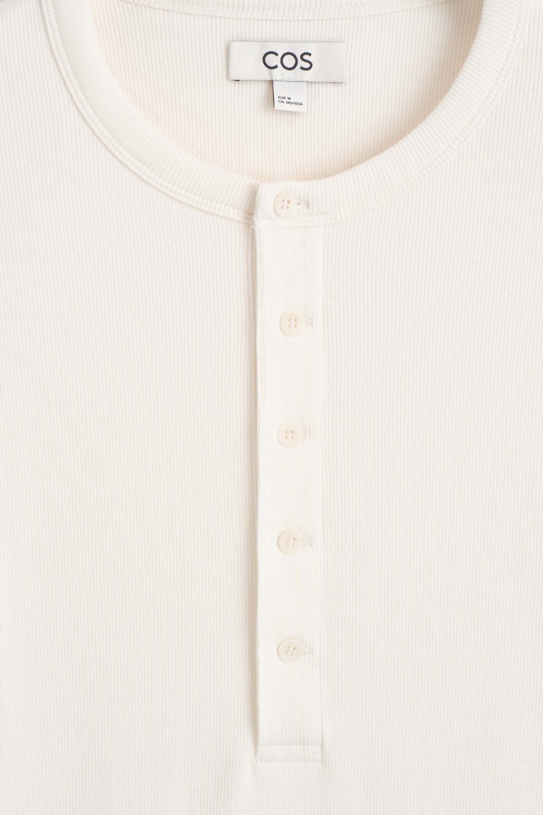 A close-up front view of the slim, ribbed cotton long-sleeved Henley T-shirt displaying its cream-coloured, finely ribbed fabric. The T-shirt features a round crew neckline with ribbed trim and a central vertical placket with five matching buttons. A close-up front view of the slim, ribbed cotton long-sleeved Henley T-shirt displaying its cream-coloured, finely ribbed fabric. The T-shirt features a round crew neckline with ribbed trim and a central vertical placket with five matching buttons.