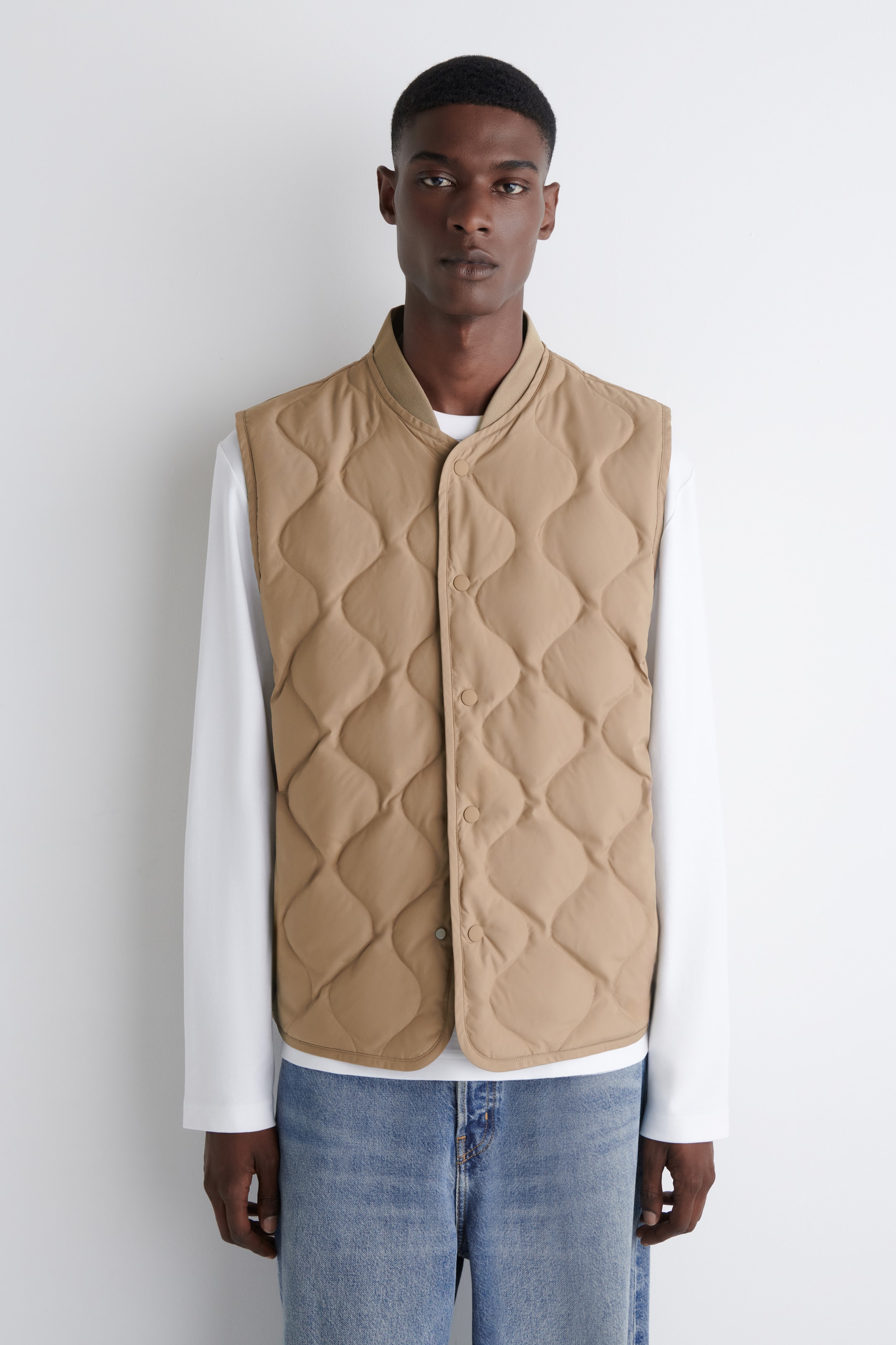 Agrandir l'image: Un gilet matelassé beige est présenté, avec un motif matelassé ondulé et un col à bande en tricot côtelé. Il présente une fermeture à boutons-pression sur toute la longueur et est porté par-dessus une chemise blanche à manches longues et un jean bleu.