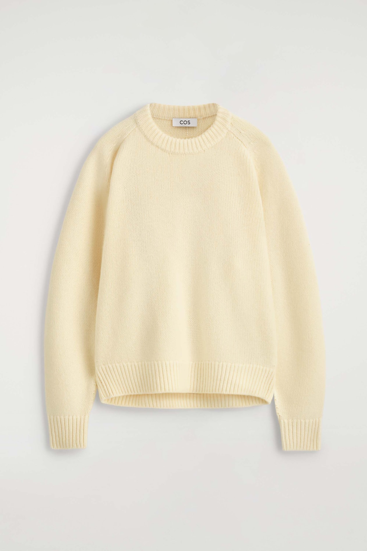 A cream-coloured rounded cashmere crew-neck jumper is shown from the front, featuring a ribbed crew neckline, raglan sleeves, and a soft knit texture. The jumper showcases ribbed cuffs and a matching ribbed hem that define its relaxed silhouette.