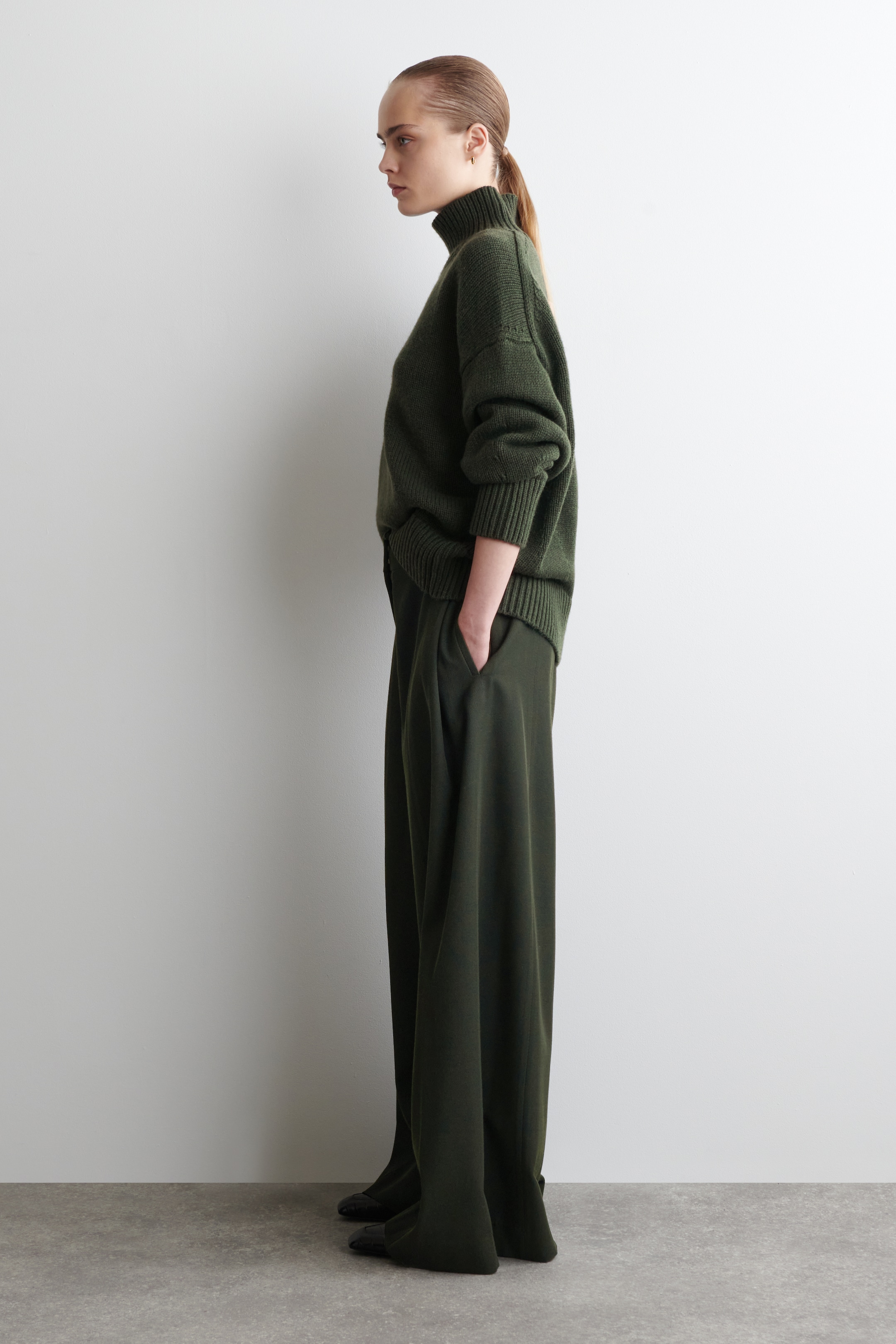 Größeres Bild anzeigen: Ein dunkelgrüner Rollkragenpullover und passende weite Hosen werden aus einer Seitenansicht präsentiert. Der locker sitzende Pullover zeichnet sich durch einen hohen gerippten Kragen, gerippte Bündchen und einen gerippten Saum aus, während die weiten Hosen eine lockere, fließende Silhouette mit einer Hand, die in einer Seitentasche ruht, zeigen.
