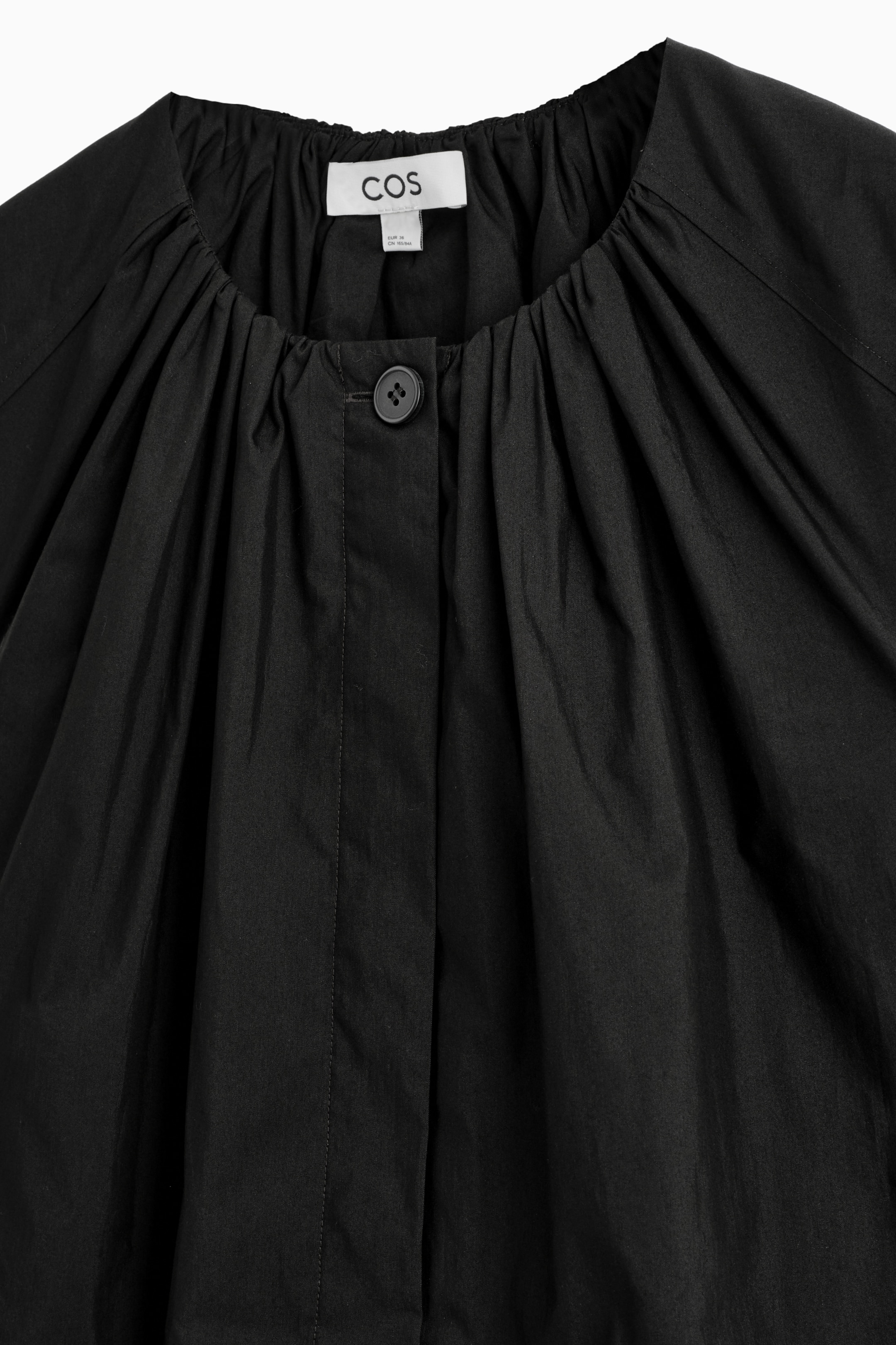 Agrandir l'image: Le devant d'une robe noire présente un col rond haut et froncé avec une fermeture à bouton unique. Le tissu tombe de manière volumineuse depuis le col.