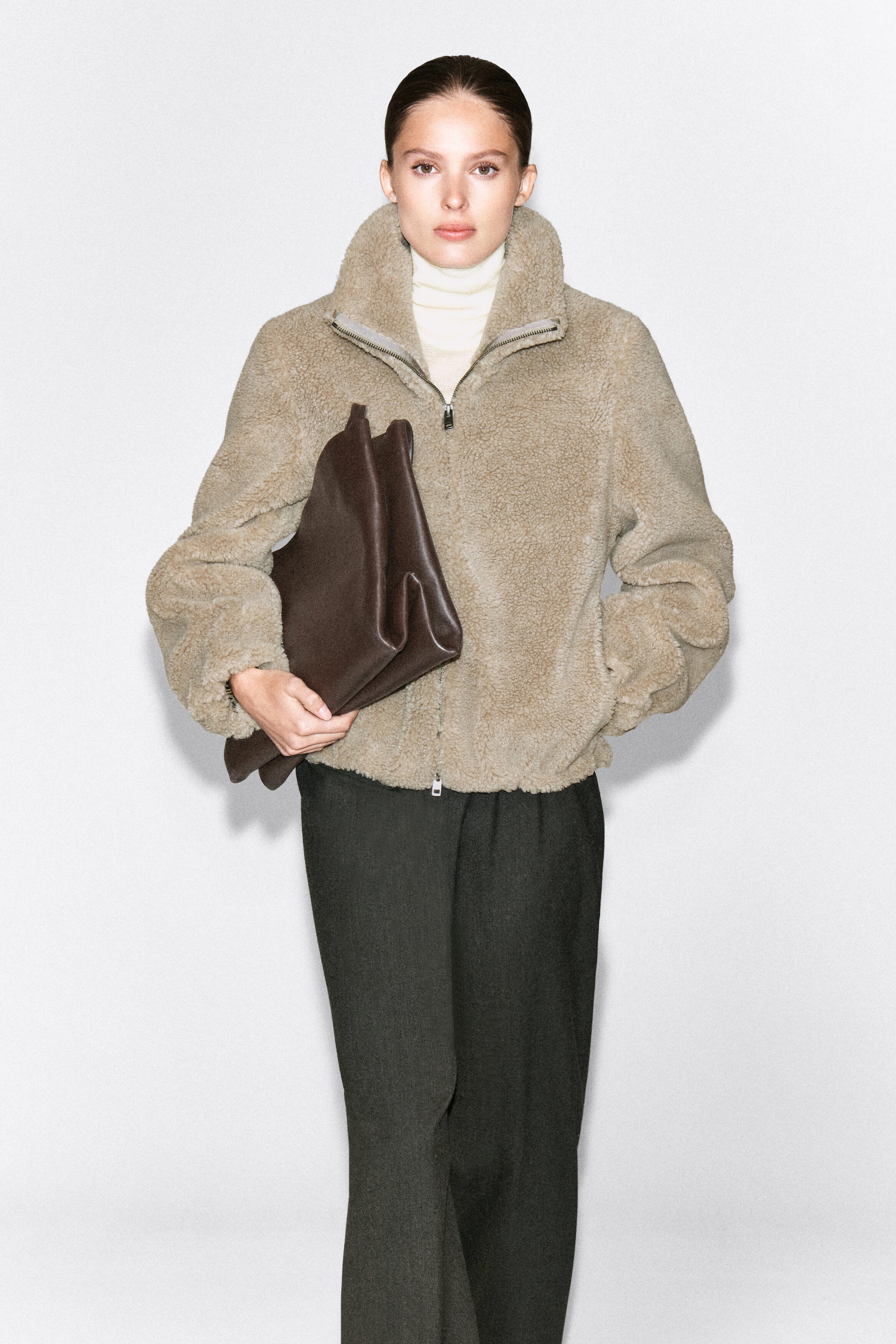 A beige funnel-neck teddy jacket in textured fleece is being worn by the model, featuring a front zip closure and side pockets. The jacket is layered over a cream turtleneck top and styled with dark trousers, with the model holding a dark brown leather clutch bag.