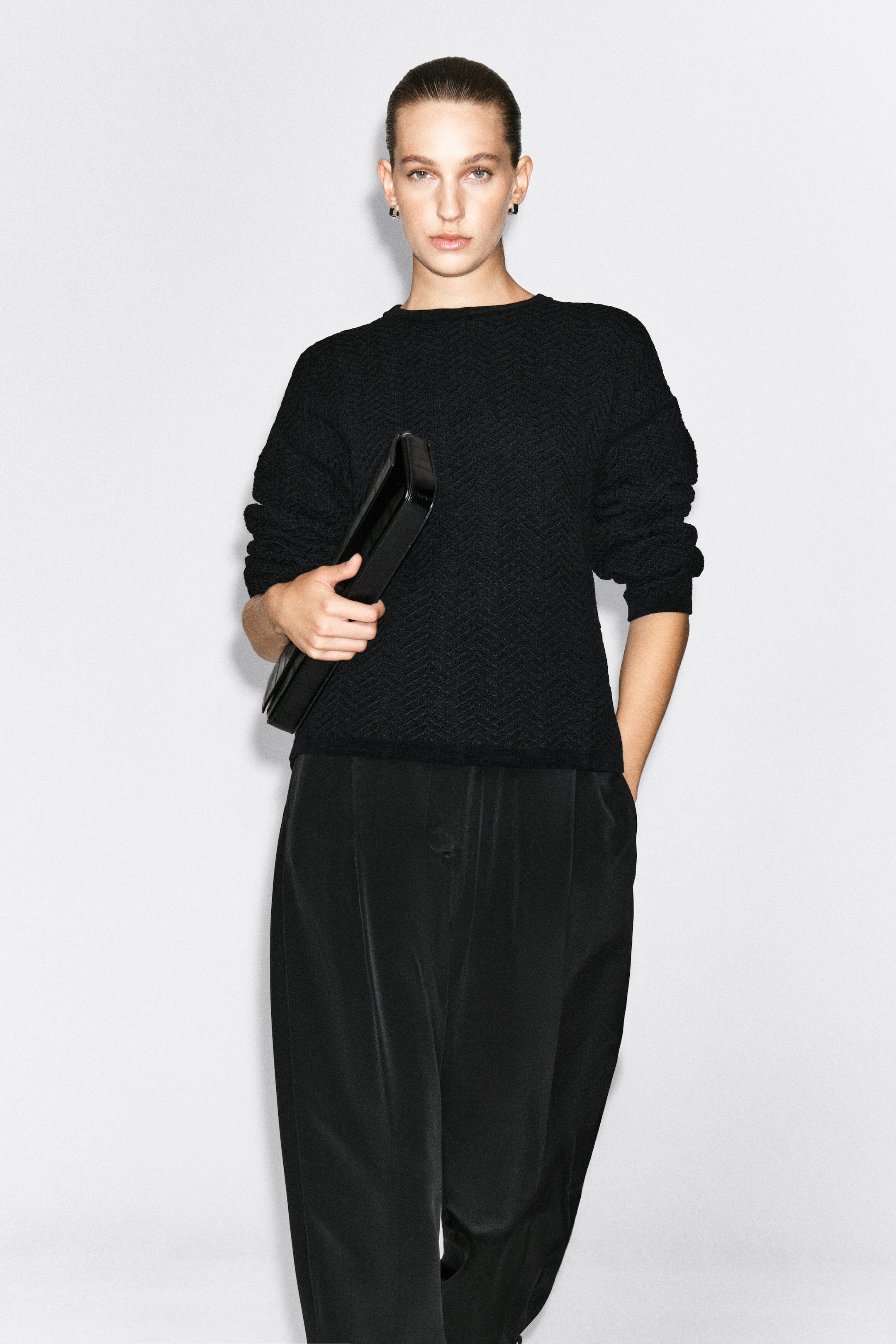 This black merino wool chenille crew-neck sweater, featuring a textured chevron knit pattern, is worn by the model. The sweater is styled with loose black trousers, and the model is holding a black clutch bag under one arm. This black merino wool chenille crew-neck sweater, featuring a textured chevron knit pattern, is worn by the model. The sweater is styled with loose black trousers, and the model is holding a black clutch bag under one arm.