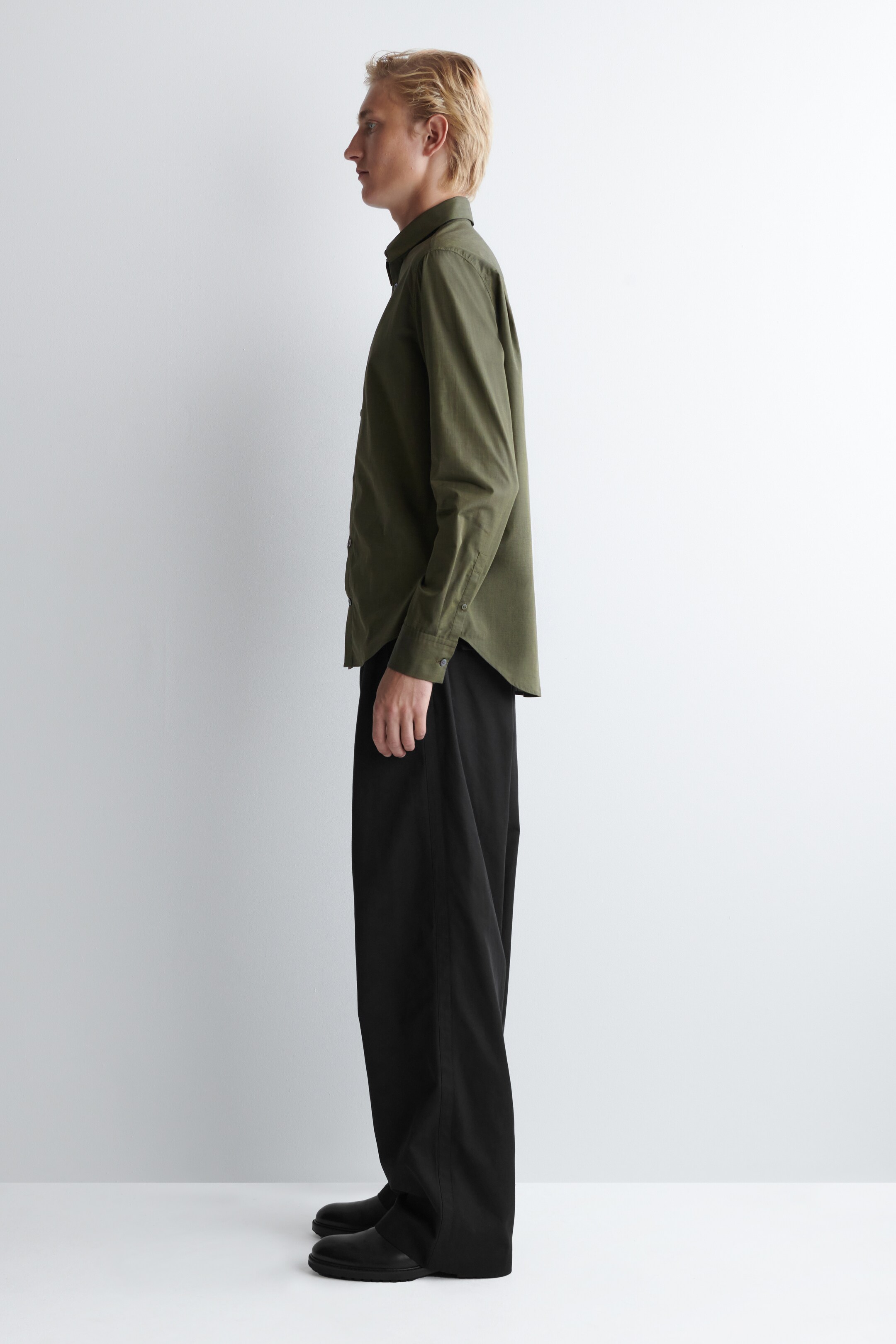Ver imagen más grande: Una camisa de manga larga verde oliva se está usando, vista de lado, mostrando un cuello clásico y puños con botones. La camisa está metida en pantalones anchos negros que caen sueltos sobre zapatos negros.