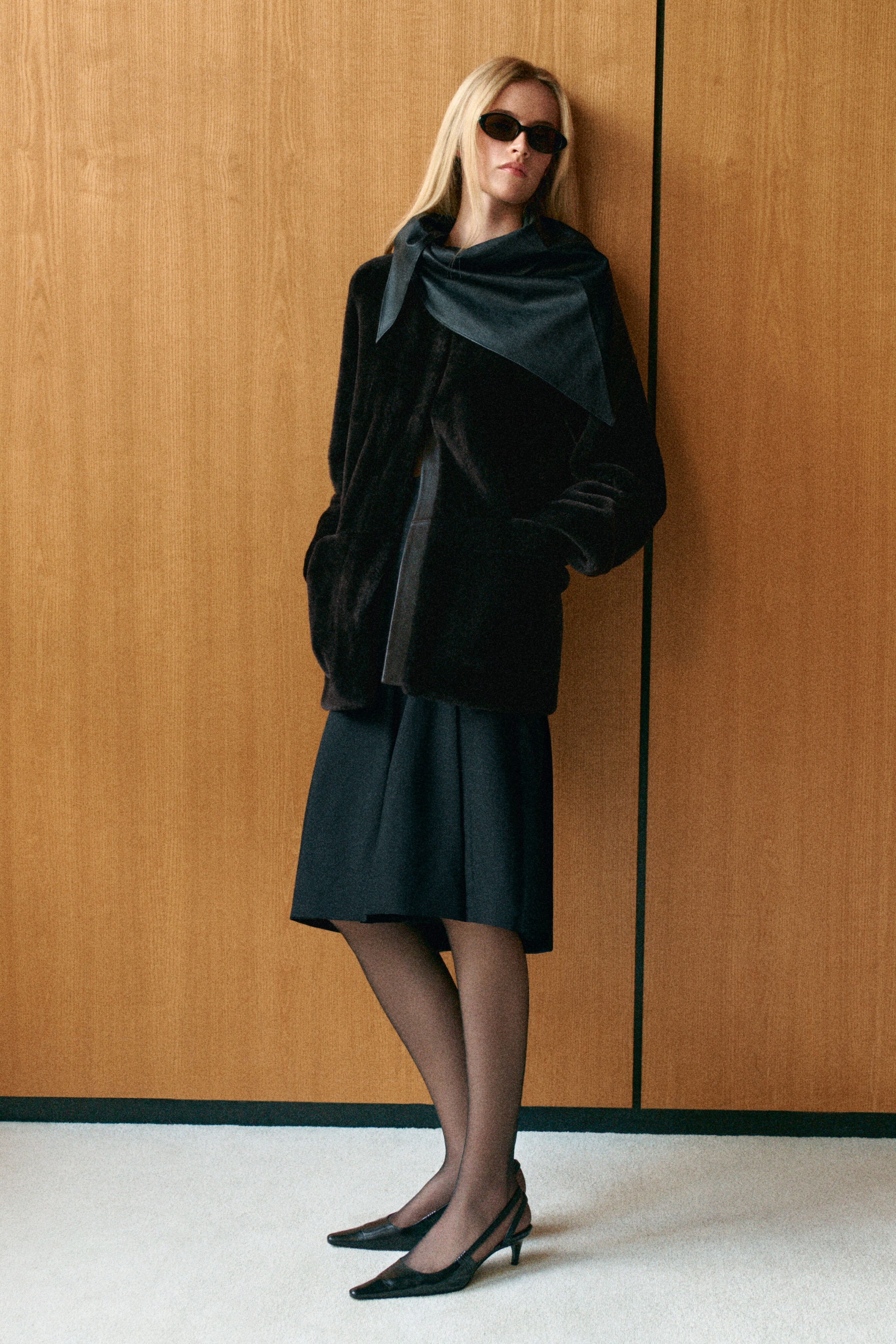 A collarless shearling jacket in dark brown is shown from the front, featuring a plush, textured finish and a relaxed, hip-length silhouette. The jacket is worn open with hands resting in the side pockets, and is styled with a matching dark scarf draped over the collarless neckline. A collarless shearling jacket in dark brown is shown from the front, featuring a plush, textured finish and a relaxed, hip-length silhouette. The jacket is worn open with hands resting in the side pockets, and is styled with a matching dark scarf draped over the collarless neckline.