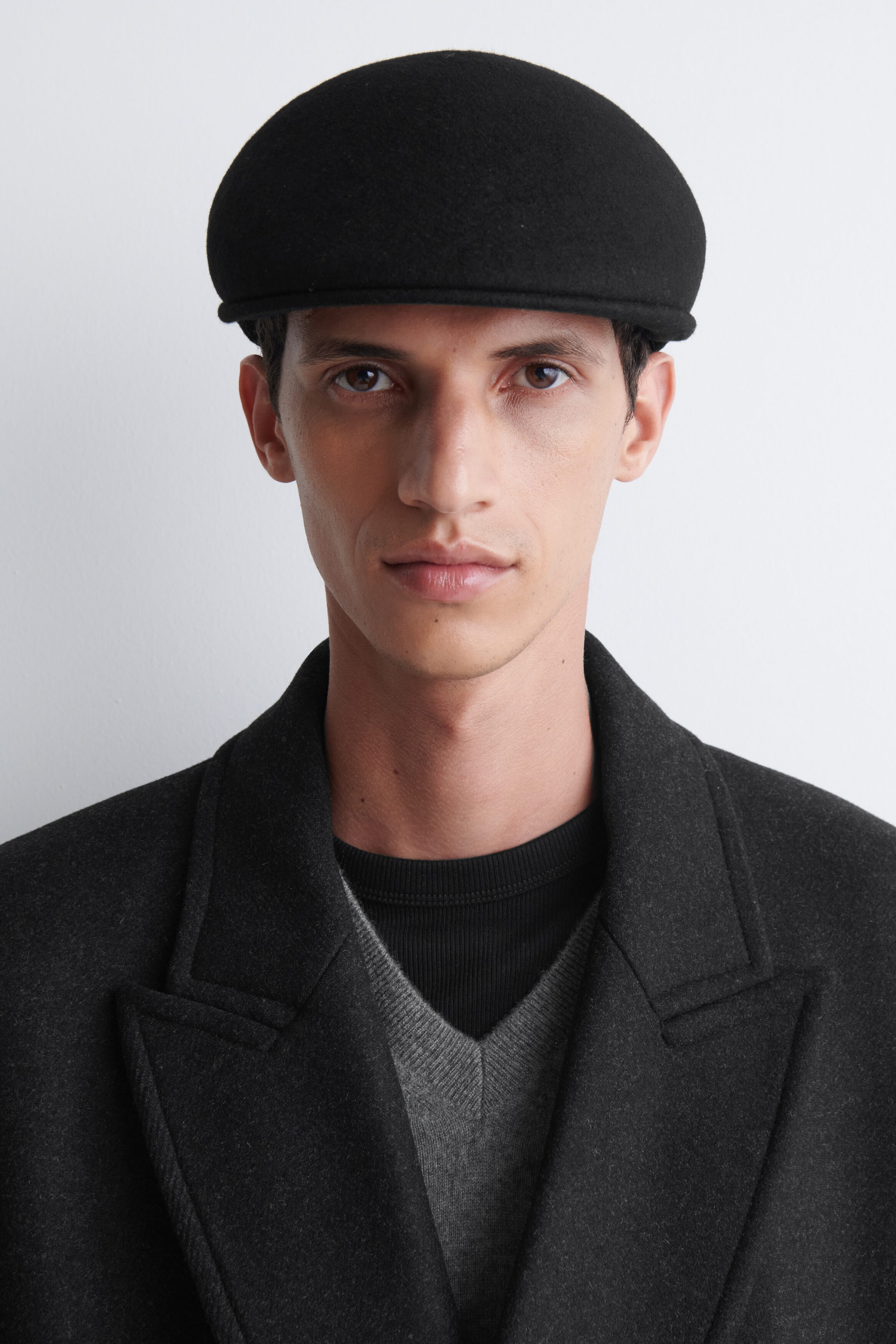 Le mannequin porte une casquette plate noire à la silhouette arrondie et structurée. La casquette est mise en valeur avec un manteau en laine gris foncé superposé à un pull gris à col en V. Le mannequin porte une casquette plate noire à la silhouette arrondie et structurée. La casquette est mise en valeur avec un manteau en laine gris foncé superposé à un pull gris à col en V.