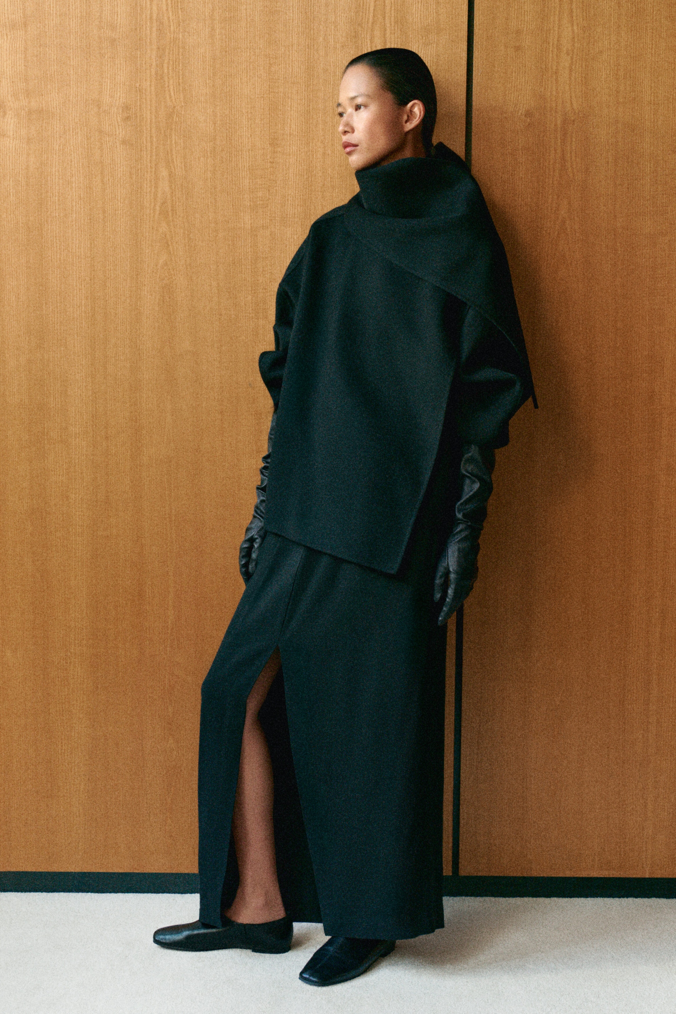 A black double-faced wool-blend scarf jacket presents a relaxed yet structured silhouette, featuring an integrated draped scarf wrapped around the neck. Styled from a front-facing angle, it is paired with long black leather gloves and a matching black maxi skirt with a front slit.