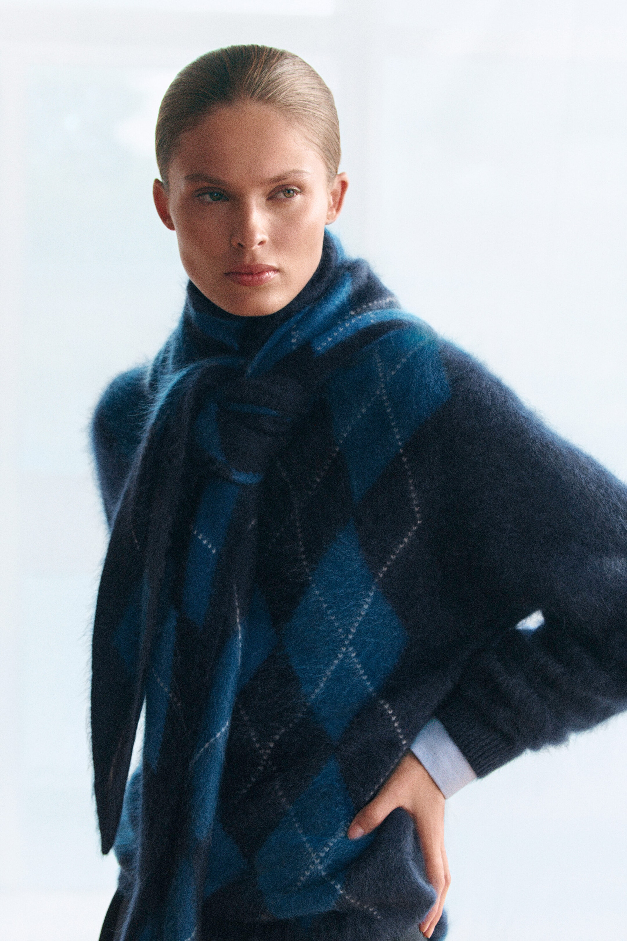 A navy and teal argyle jumper featuring a brushed, fuzzy texture is shown from a three-quarter front view on a model. The jumper is styled with a matching argyle scarf tied around the neck and features ribbed cuffs with a light blue sleeve peeking out at the wrist. A navy and teal argyle jumper featuring a brushed, fuzzy texture is shown from a three-quarter front view on a model. The jumper is styled with a matching argyle scarf tied around the neck and features ribbed cuffs with a light blue sleeve peeking out at the wrist.