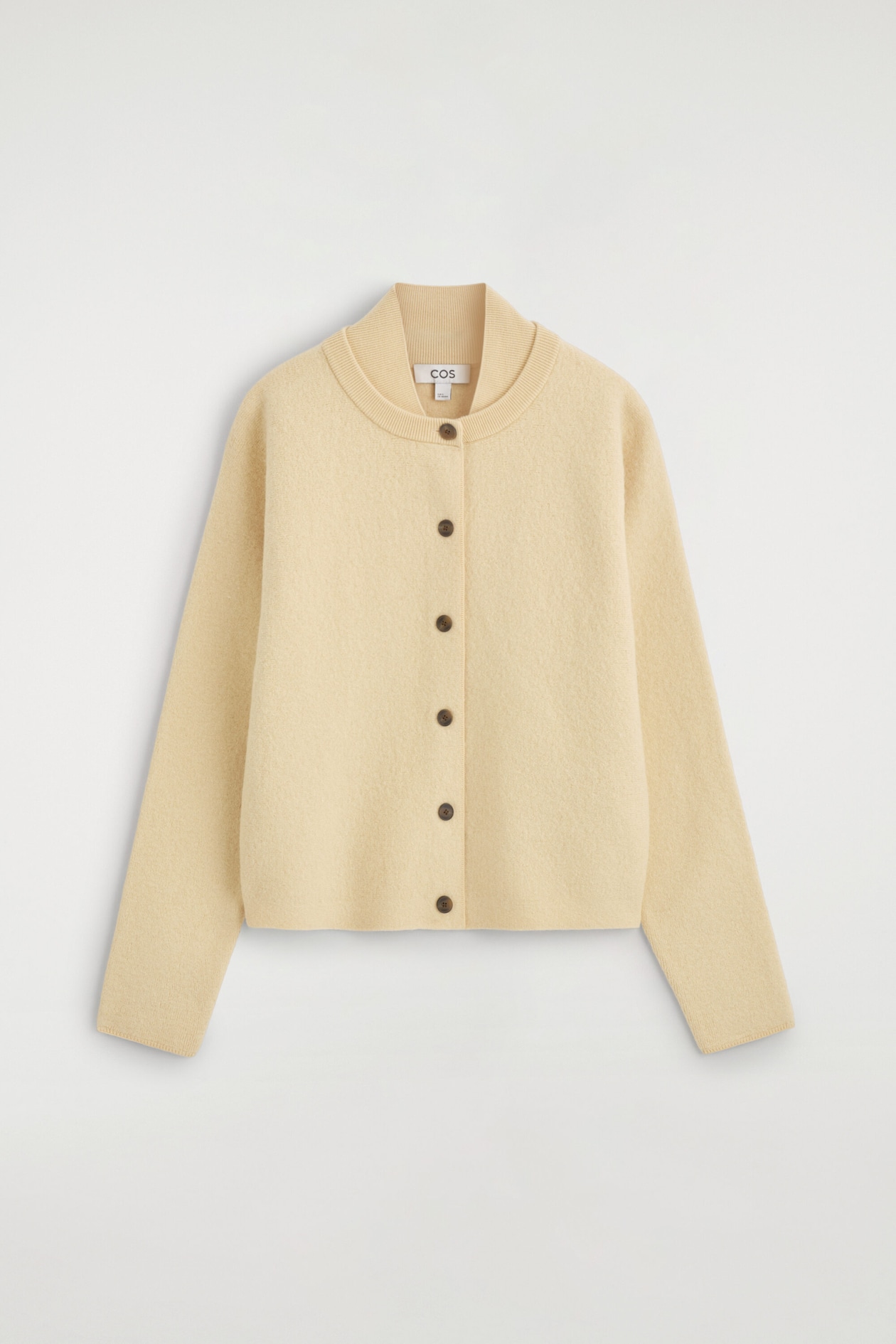 Un cardigan en laine bouillie jaune pâle est présenté de face, avec une silhouette doucement arrondie et un col côtelé unique à double épaisseur. Le cardigan se ferme avec six boutons brun foncé au centre devant et présente de longues manches légèrement fuselées avec des poignets aux finitions nettes.