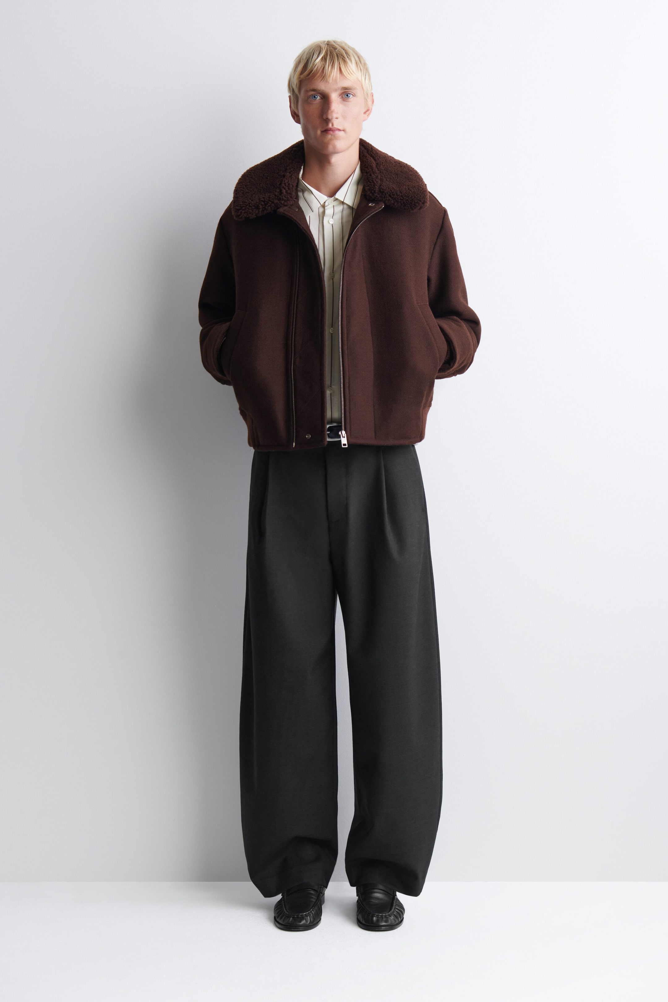 The dark brown wool jacket is shown facing forward, featuring a plush, textured shearling collar, a silver zip-front closure, and a relaxed, boxy fit. It is styled over a light-colored vertical-striped shirt and wide-leg, pleated black trousers. The dark brown wool jacket is shown facing forward, featuring a plush, textured shearling collar, a silver zip-front closure, and a relaxed, boxy fit. It is styled over a light-colored vertical-striped shirt and wide-leg, pleated black trousers.