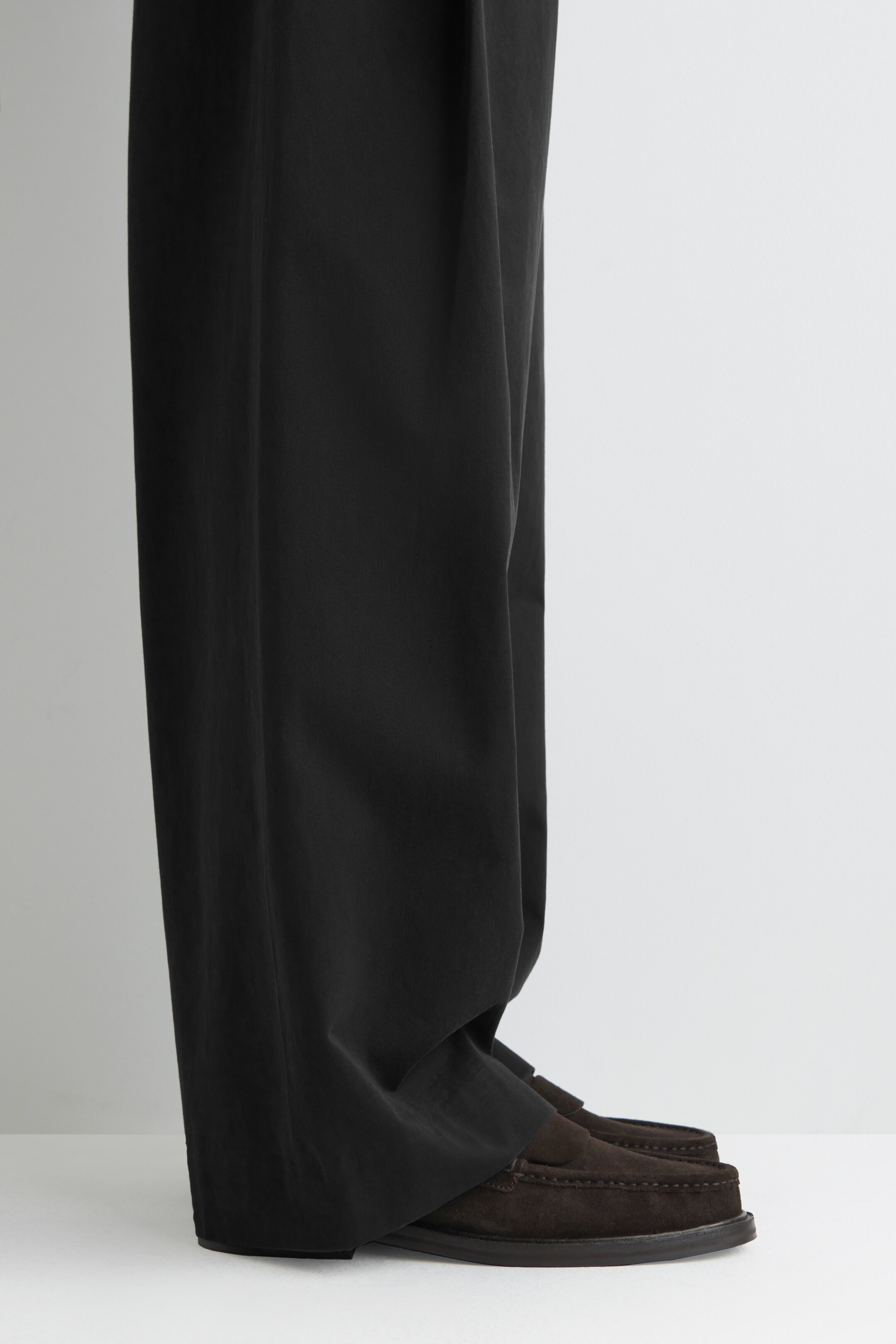 Agrandir l'image: Un pantalon noir à jambes larges tombe librement, couvrant la partie supérieure d'une paire de mocassins en daim marron. Le pantalon présente un drapé décontracté lorsqu'il rencontre les chaussures, qui sont vues de côté.