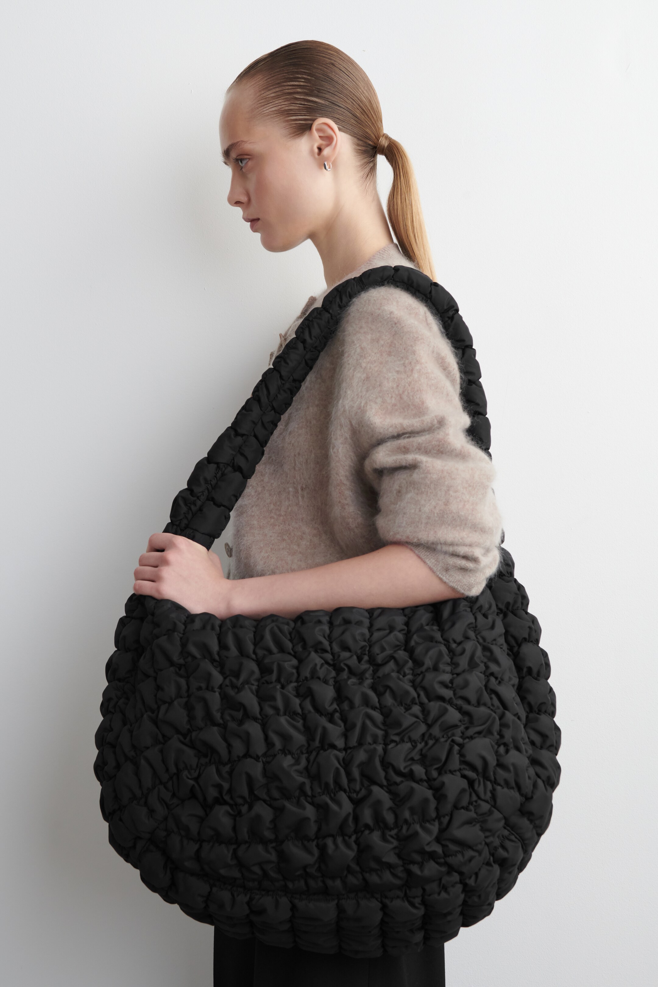 Visualizza immagine più grande: Una grande borsa a tracolla nera viene indossata lateralmente, con il suo corpo arrotondato e la tracolla larga caratterizzati da una texture voluminosa e arricciata distintiva.