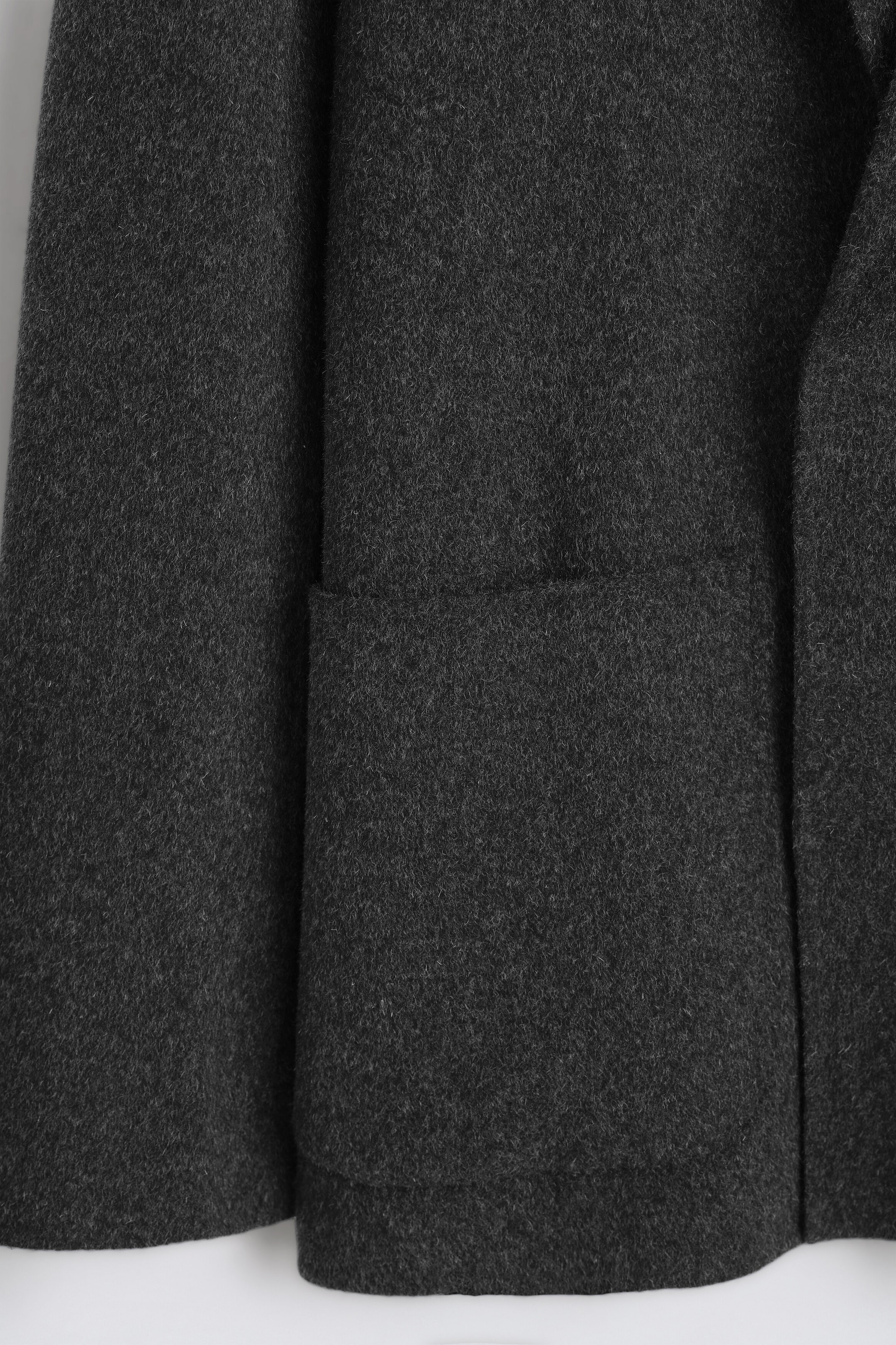 A close-up view of a dark grey wool-blend blazer highlighting the soft, textured surface of the double-faced fabric, showing a large patch pocket and the clean finish of the bottom hem. A close-up view of a dark grey wool-blend blazer highlighting the soft, textured surface of the double-faced fabric, showing a large patch pocket and the clean finish of the bottom hem.