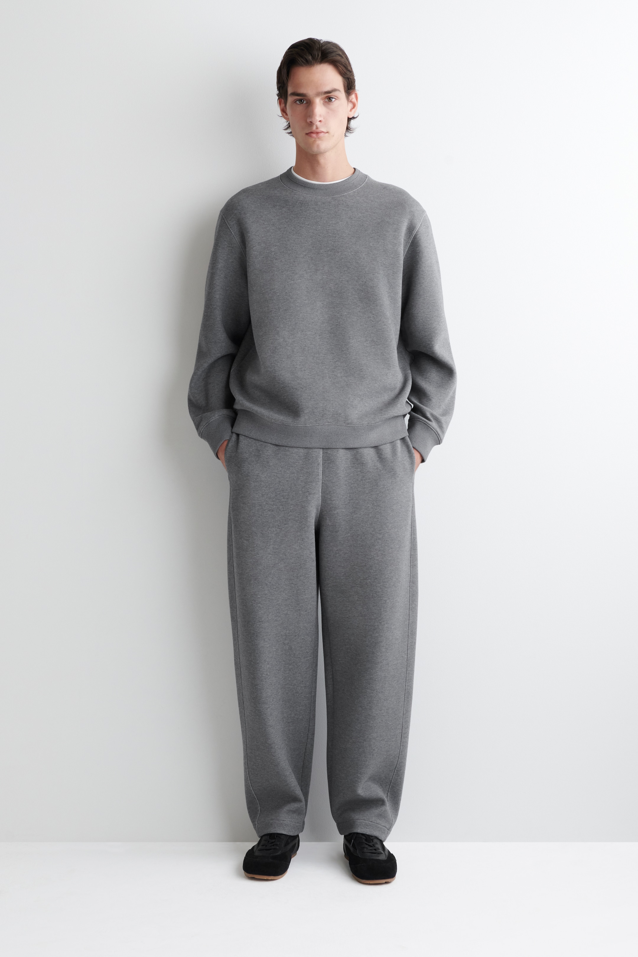 Größeres Bild ansehen: Ein locker sitzendes graues Loungewear-Set wird in einer Ganzkörperansicht von vorne präsentiert. Es verfügt über ein Sweatshirt mit Rundhalsausschnitt, gerippten Bündchen und Saum, das über einem sichtbaren weißen Rundhals-T-Shirt getragen wird, und passende Jogginghosen mit einem elastischen Bund und geraden Säumen.
