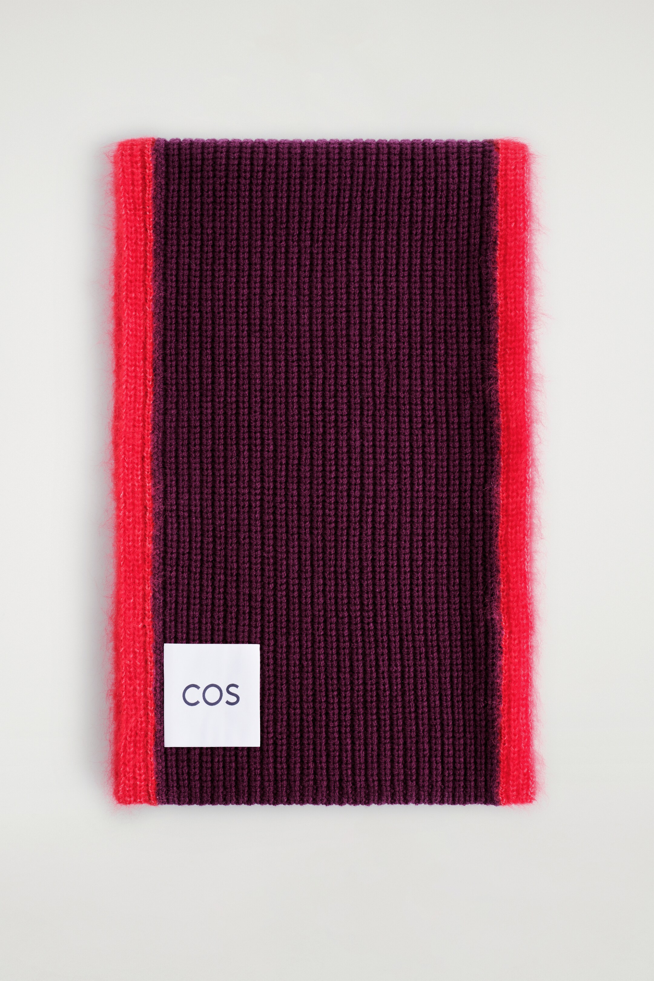 Agrandir l'image: Une écharpe en maille côtelée est pliée, affichant une section principale prune profond avec des bords verticaux rouges vibrants et flous. Une étiquette blanche "COS" est positionnée sur la zone prune.
