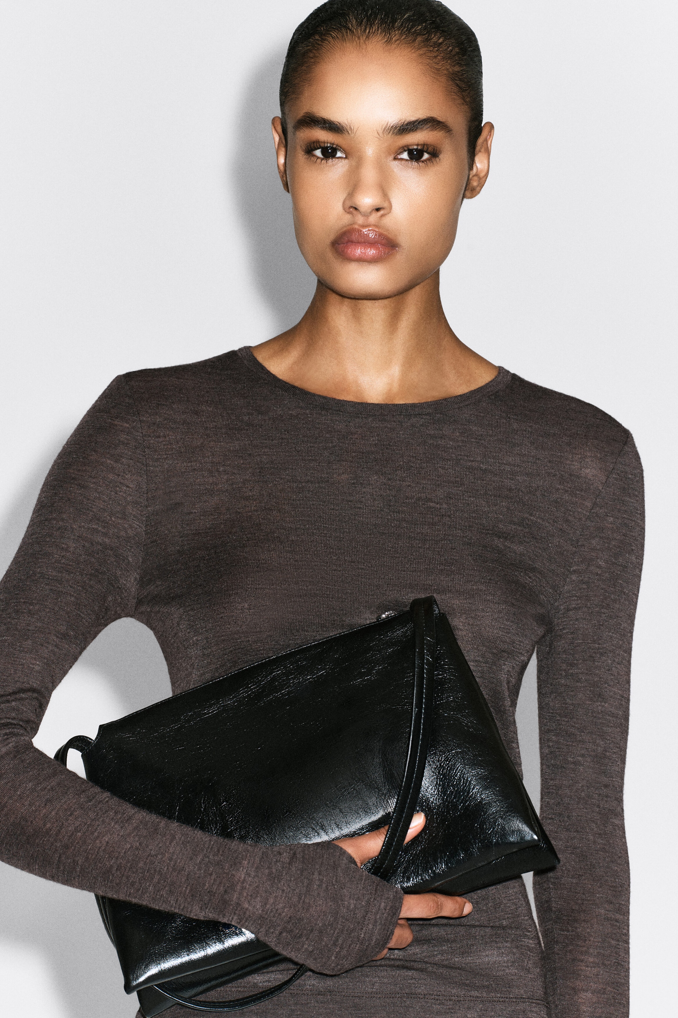 The slim merino wool top is shown from the front in a close-fitting silhouette, featuring a lightweight, heathered dark brown knit and a round neckline. The long sleeves extend over the wrists, and a black leather bag is held against the torso. The slim merino wool top is shown from the front in a close-fitting silhouette, featuring a lightweight, heathered dark brown knit and a round neckline. The long sleeves extend over the wrists, and a black leather bag is held against the torso.