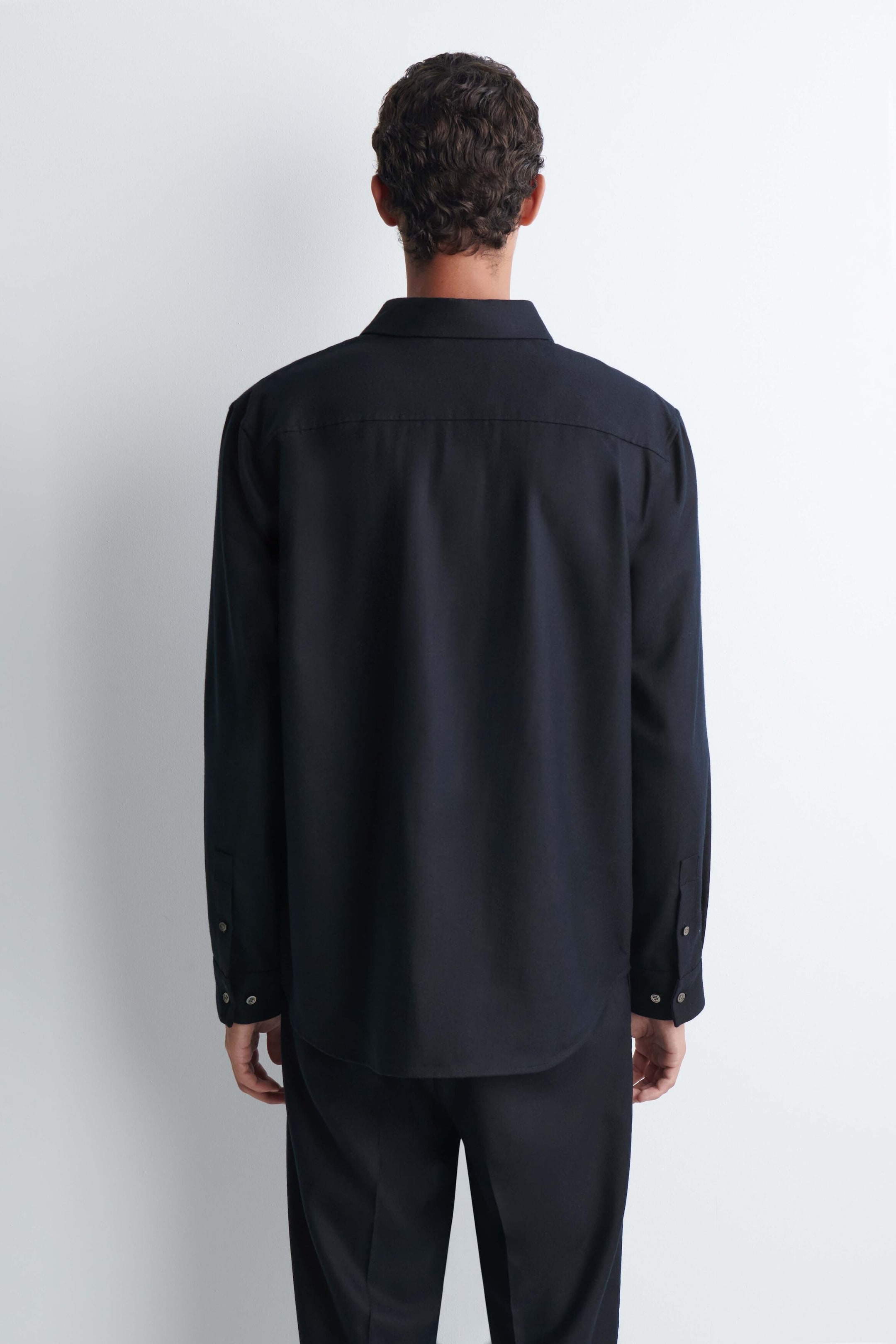 Ein dunkel marineblaues, locker geschnittenes Hemd aus Wollflanell wird vom Model getragen und zeigt die Rückansicht mit horizontaler Passe, Kragen und langen Ärmeln mit geknöpften Manschetten. Das Hemd wird mit einer passenden dunklen Hose kombiniert. Ein dunkel marineblaues, locker geschnittenes Hemd aus Wollflanell wird vom Model getragen und zeigt die Rückansicht mit horizontaler Passe, Kragen und langen Ärmeln mit geknöpften Manschetten. Das Hemd wird mit einer passenden dunklen Hose kombiniert.