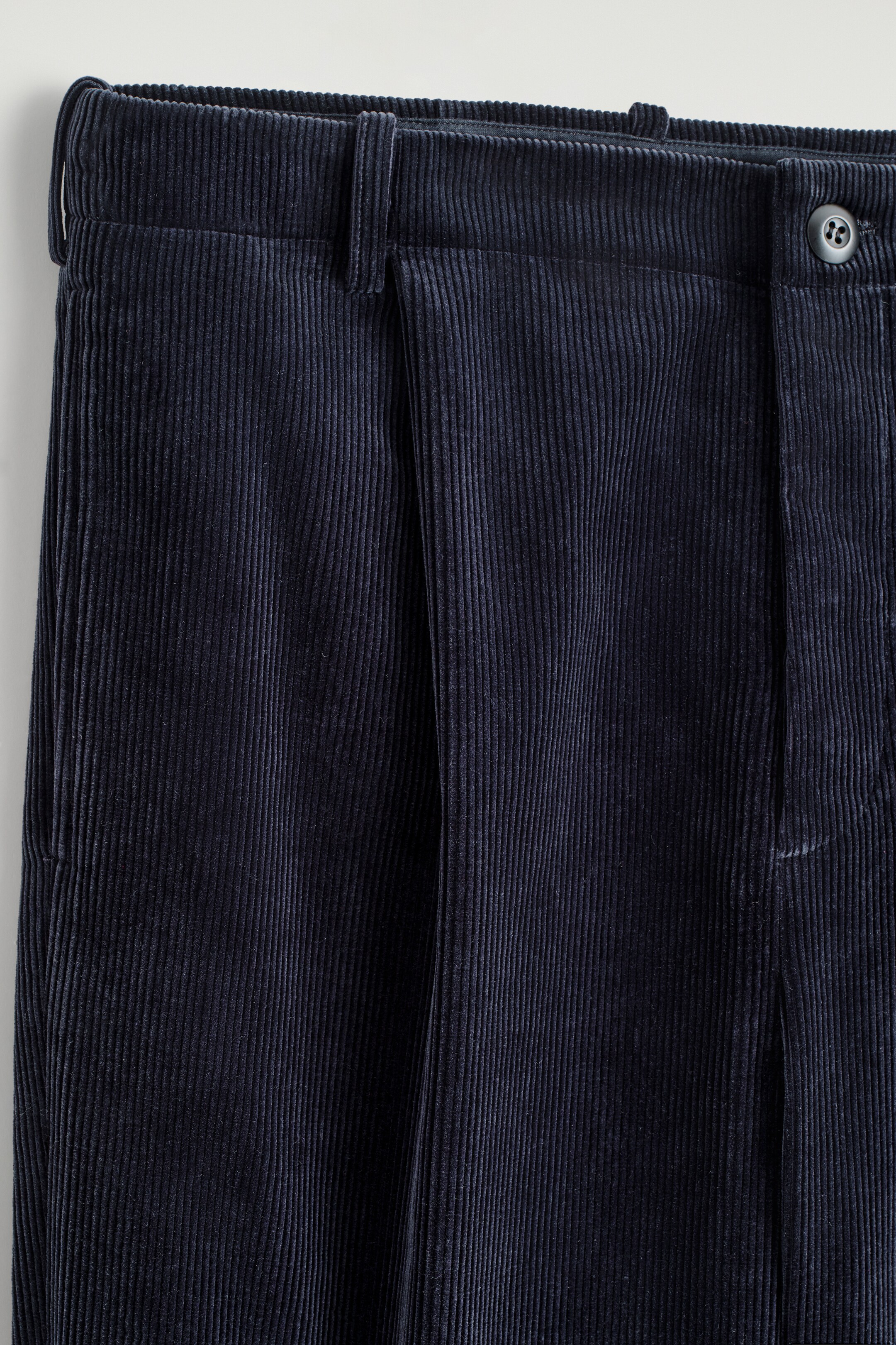 Agrandir l'image: Une paire de pantalons en velours côtelé bleu foncé met en avant ses détails de devant. Le pantalon présente une ceinture structurée avec des passants de ceinture, deux plis avant proéminents et une fermeture à bouton visible en haut à droite.
