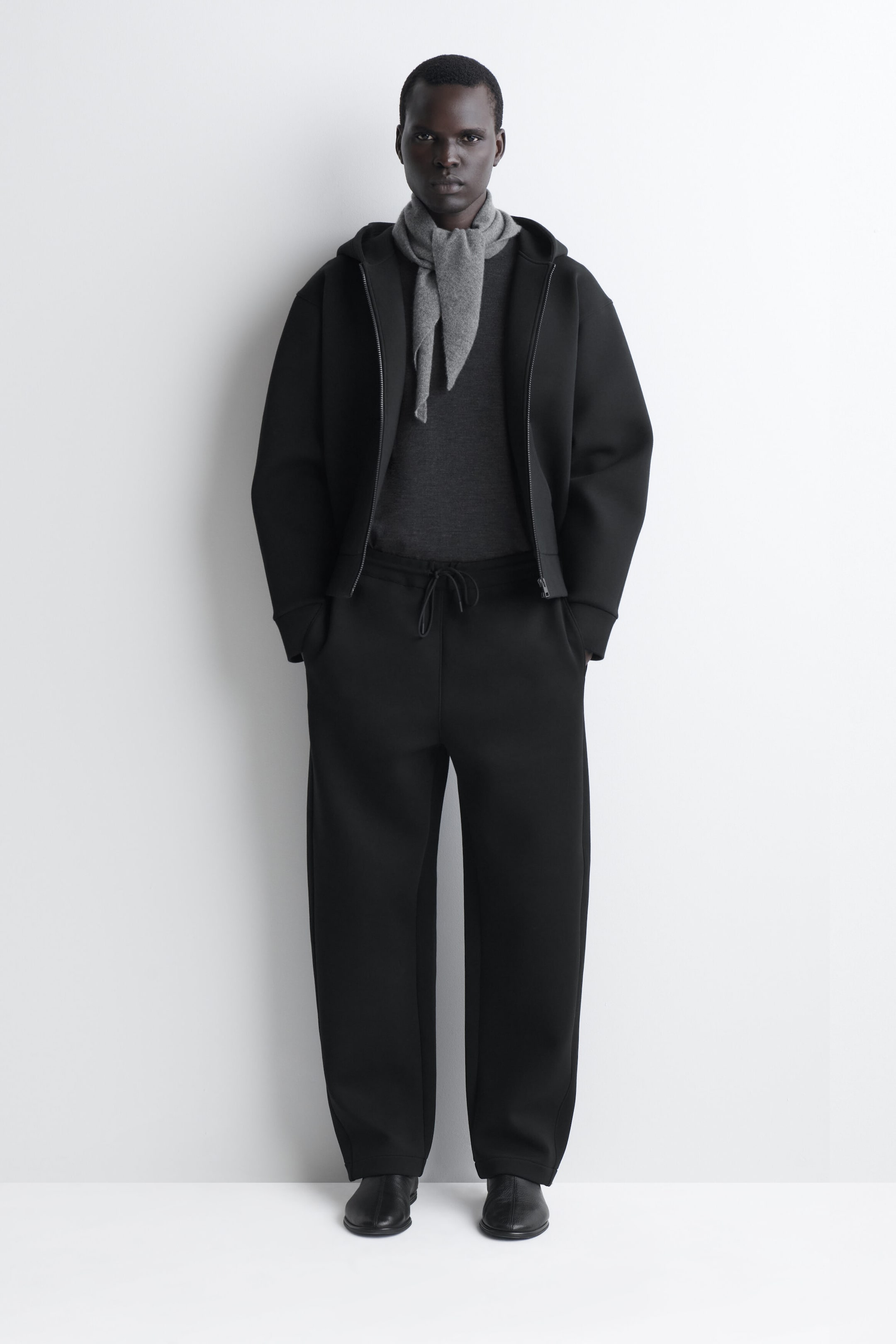 A black structured tracksuit is shown from the front, featuring an open zip-up hoodie with wide sleeves and matching relaxed-fit drawstring trousers. The set is layered over a charcoal gray knit sweater and a knotted light gray scarf, showcasing a clean, modern silhouette.