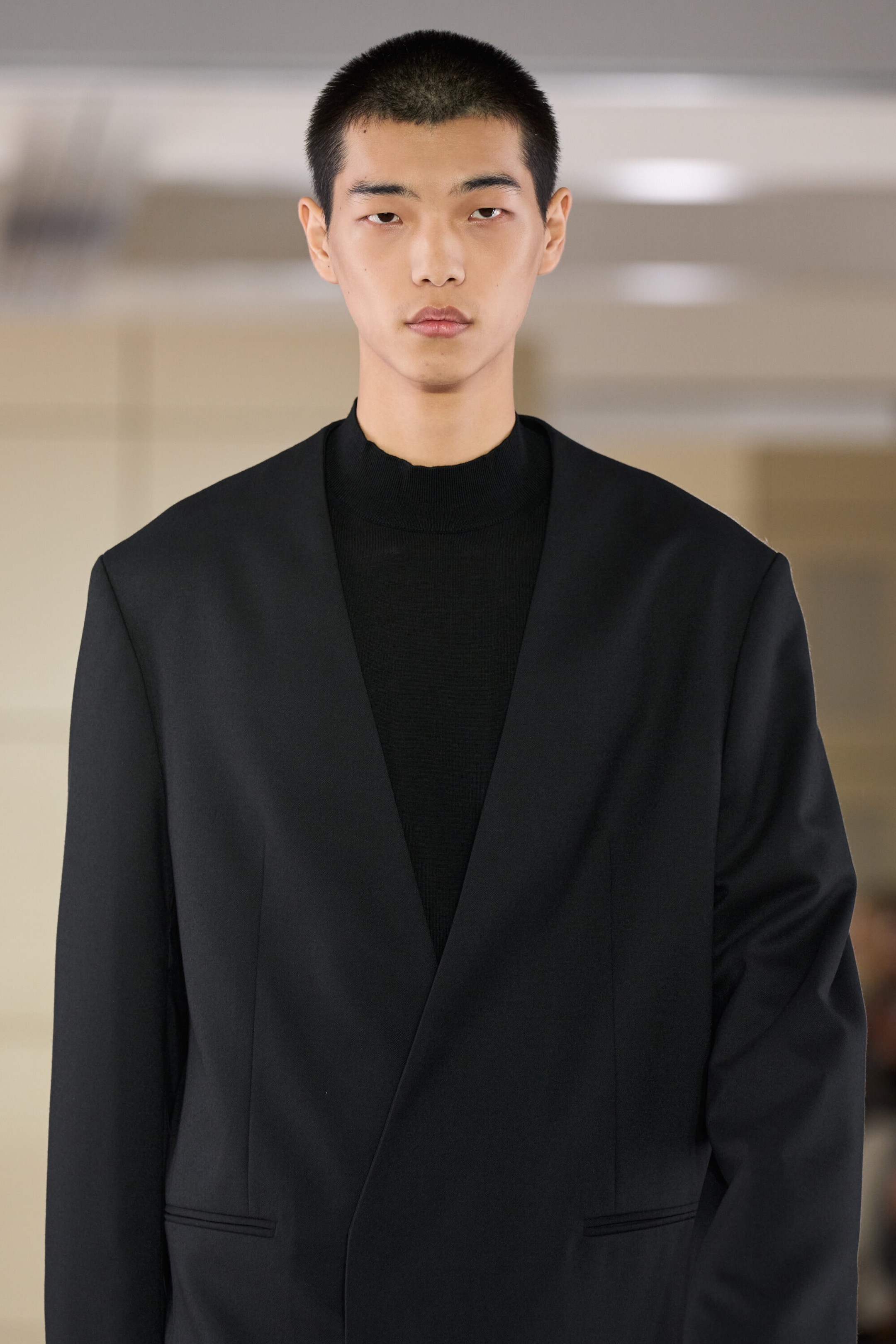 A collarless wool blazer in black is worn from the front, featuring a minimalist design with a deep V-neckline, structured shoulders, and a crossover front fastening. The blazer is layered over a black mock-neck top, highlighting its clean, streamlined fit. A collarless wool blazer in black is worn from the front, featuring a minimalist design with a deep V-neckline, structured shoulders, and a crossover front fastening. The blazer is layered over a black mock-neck top, highlighting its clean, streamlined fit.