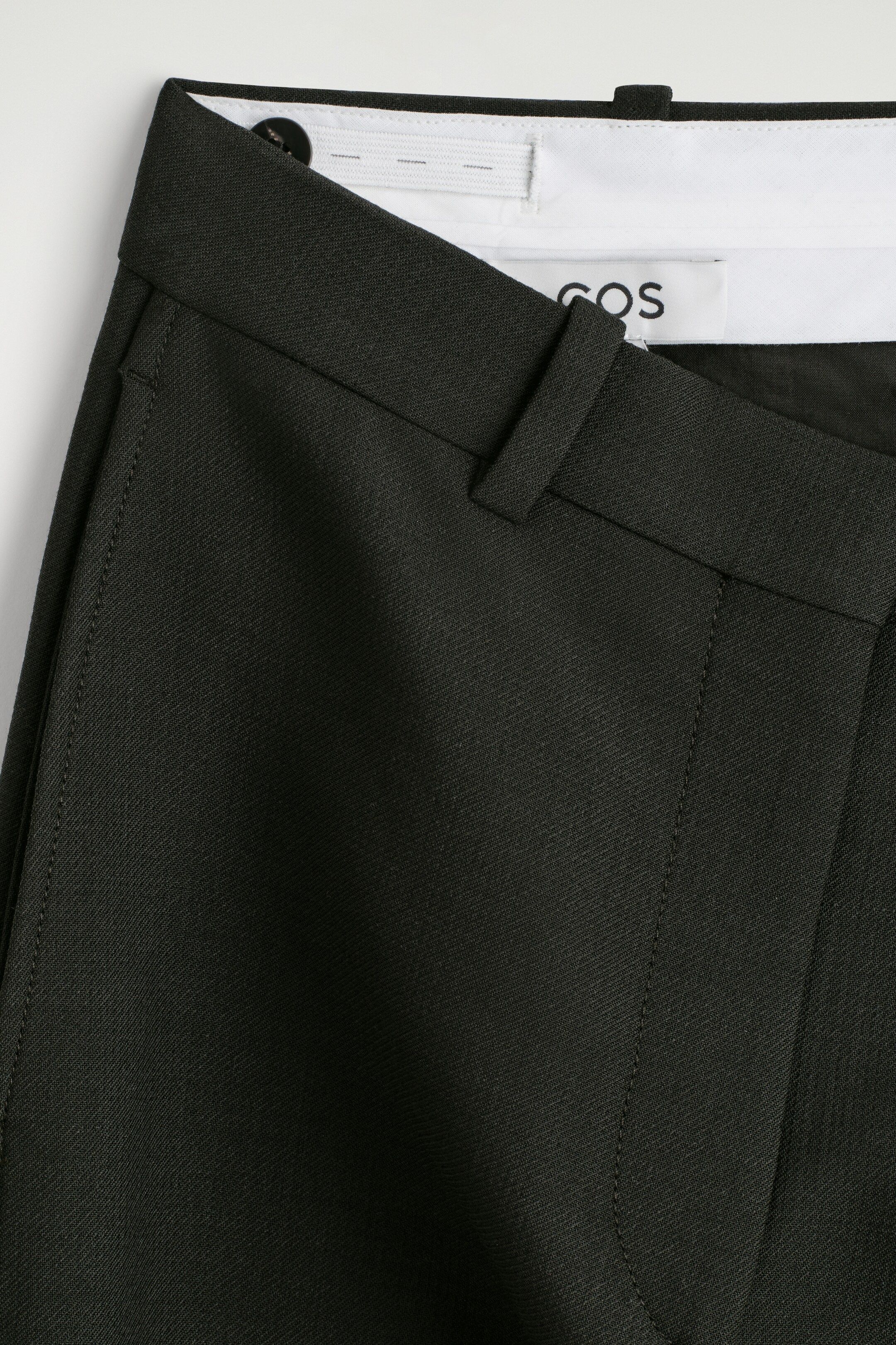 Agrandir l'image: Un pantalon de couleur taupe en tissu sergé est posé à plat, montrant une vue de FACE de la ceinture avec des passants de ceinture et des coutures incurvées vers l'avant, accentuant une silhouette arrondie. La ceinture intérieure est visiblement doublée de blanc, avec des languettes élastiques réglables et une étiquette de marque.