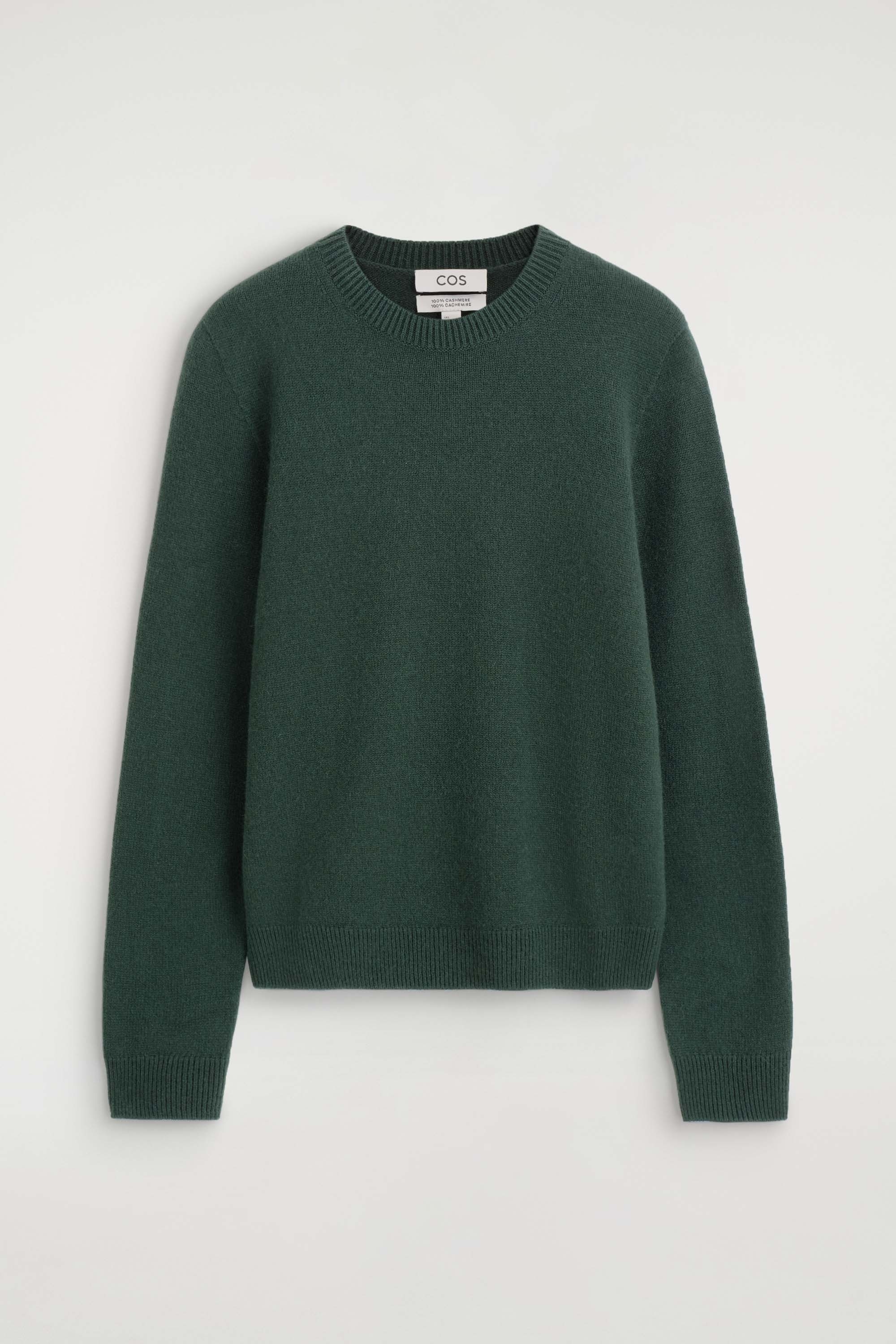 A dark green cashmere crew-neck jumper is being worn by the model, tucked into high-waisted black denim jeans. The jumper features ribbed trims at the collar and cuffs, with its sleeves slightly pushed up, and is styled with a black leather shoulder bag.