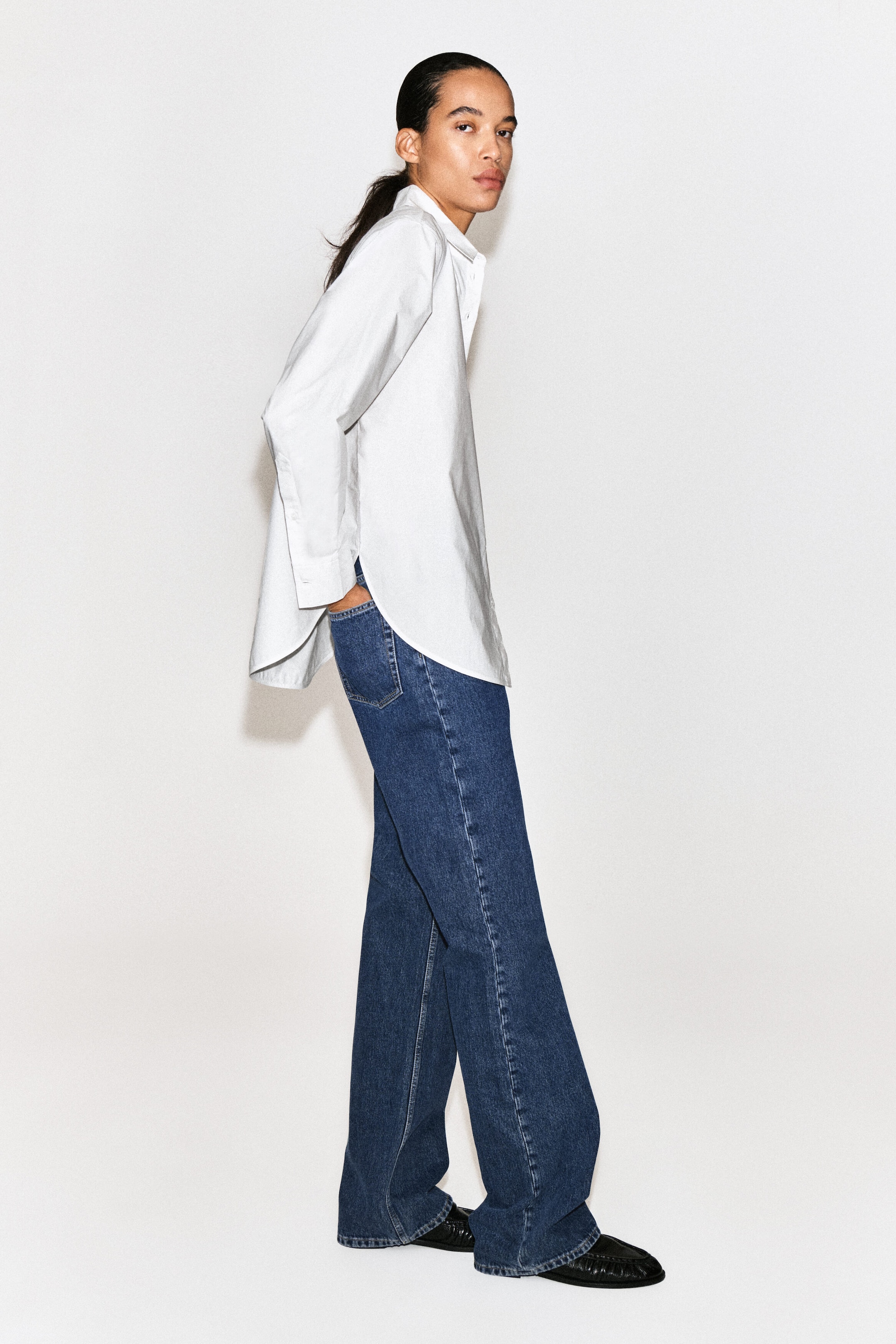 A classic cotton-poplin shirt is shown in side profile facing right, highlighting its relaxed fit, pointed collar, and long sleeves. The shirt features a curved high-low hem with deep side slits and drapes over wide-leg blue jeans. A classic cotton-poplin shirt is shown in side profile facing right, highlighting its relaxed fit, pointed collar, and long sleeves. The shirt features a curved high-low hem with deep side slits and drapes over wide-leg blue jeans.
