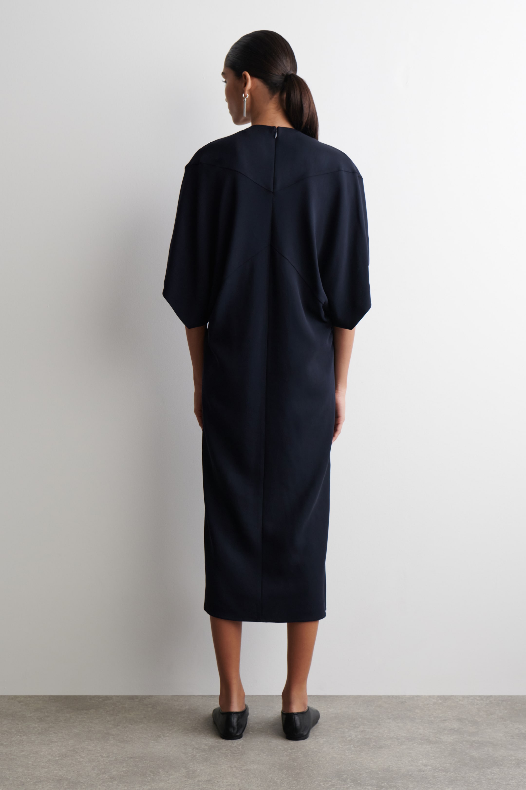 Grösseres Bild ansehen: Ein dunkelblaues Midikleid wird von hinten betrachtet und zeigt seine entspannte, säulenförmige Silhouette. Es verfügt über weite, dolmanartige Ärmel, einen hohen Kragen mit einem sichtbaren Reißverschluss in der hinteren Mitte und markante V-förmige Nähte über dem oberen Rücken.