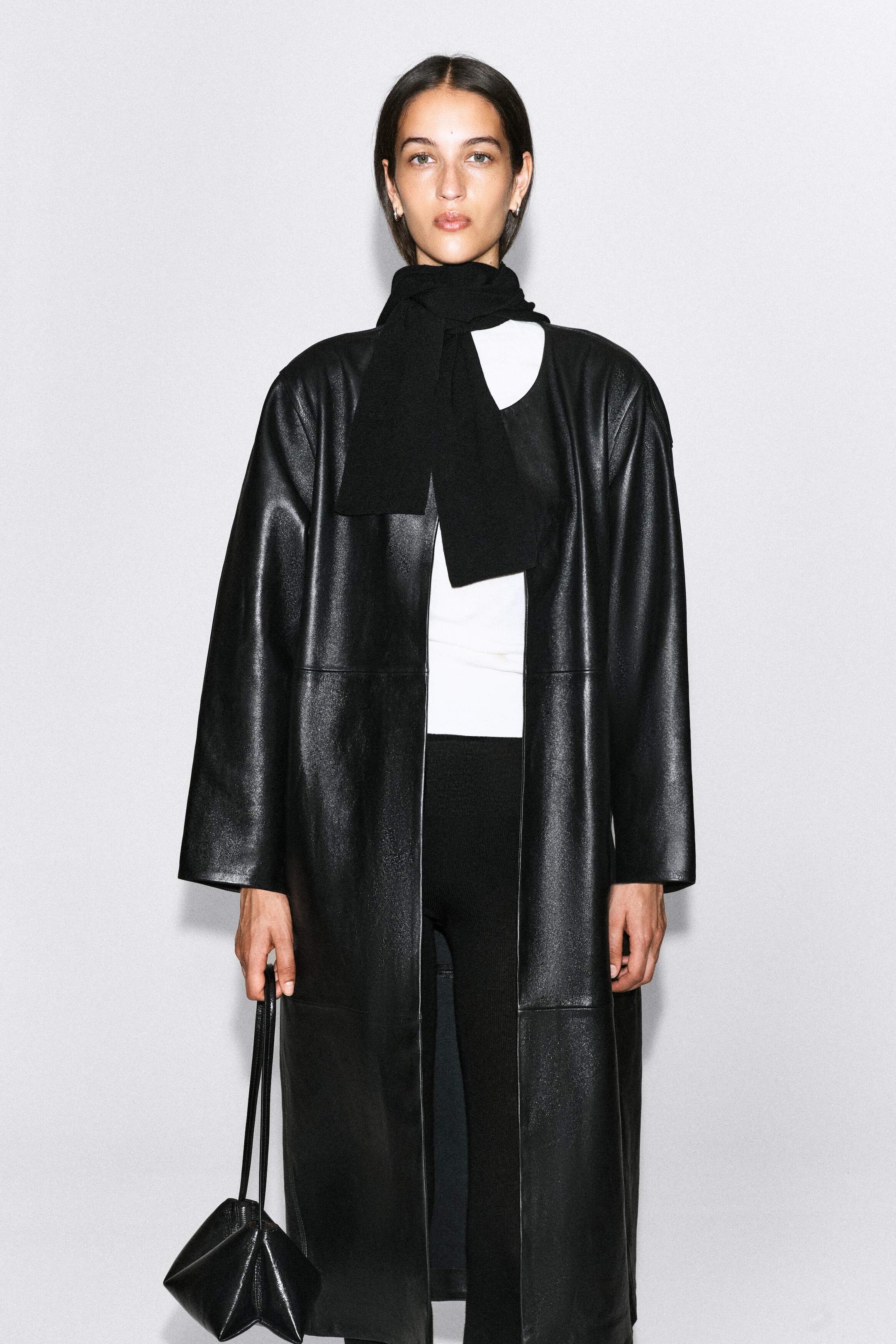 A black knitted hooded scarf is worn wrapped around the model's neck, with its ends hanging down the front over a white top and a long black leather coat. The model is also carrying a black leather handbag. A black knitted hooded scarf is worn wrapped around the model's neck, with its ends hanging down the front over a white top and a long black leather coat. The model is also carrying a black leather handbag.