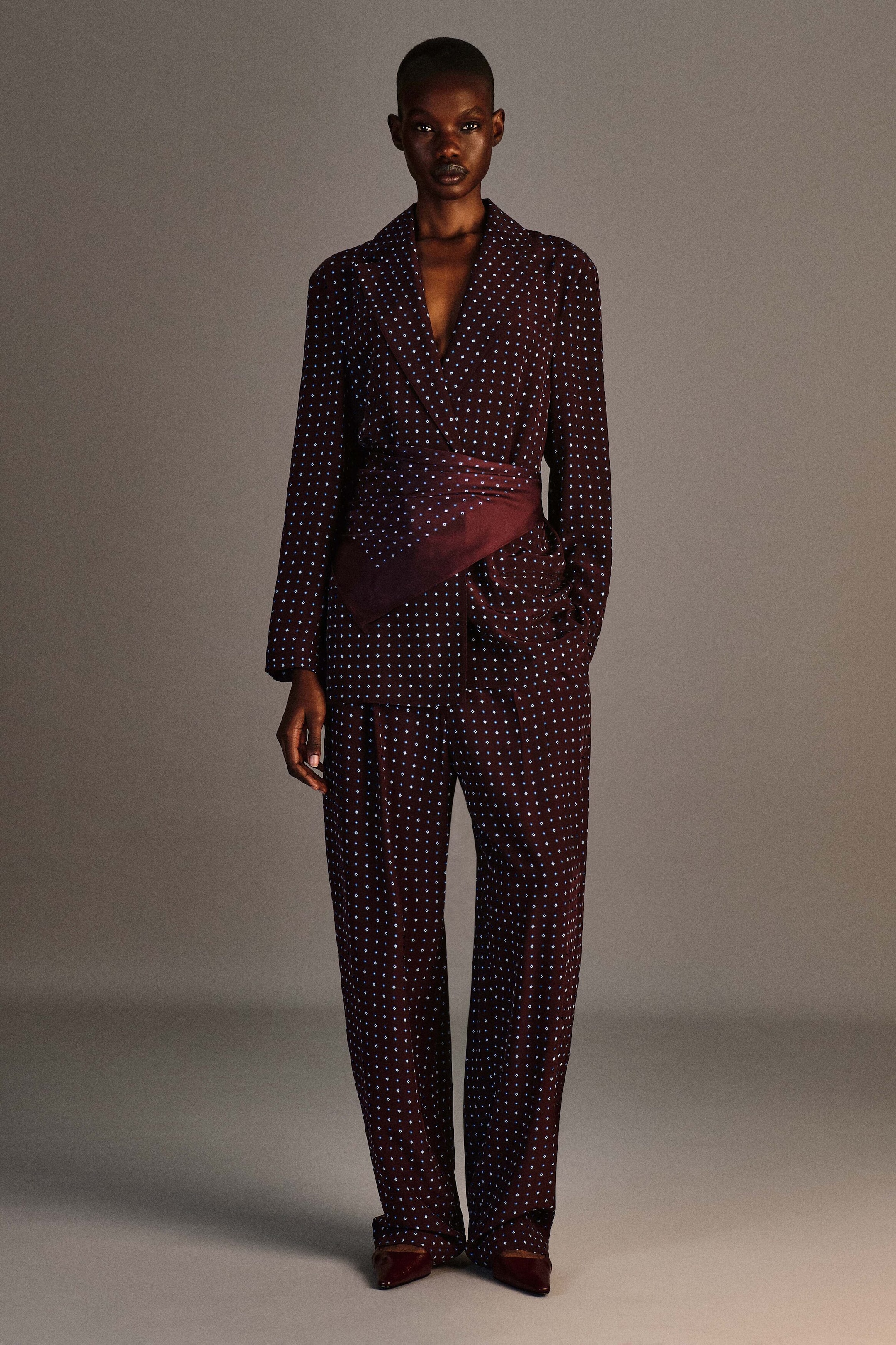 Un mannequin est de face, portant un blazer croisé à motif losange d’un bordeaux profond, doté de revers crantés et d’une large écharpe drapée assortie enroulée autour de la taille. Le blazer est associé à un pantalon droit assorti à motif losange qui tombe jusqu’à laisser apparaître le bout de chaussures bordeaux à bout pointu.