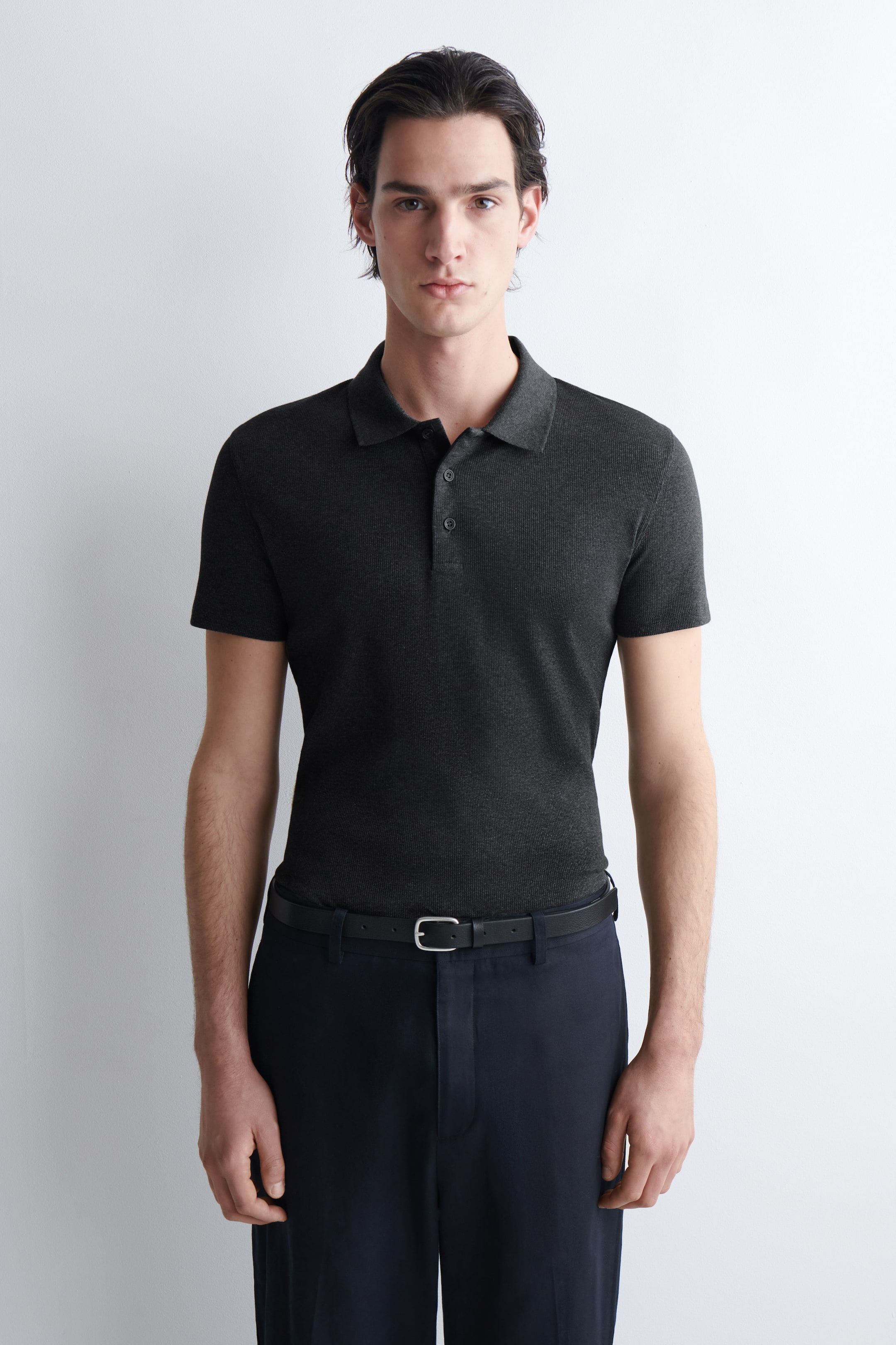 View larger image: A dark-grey polo shirt is presented facing forwards, featuring a subtle textured knit, a classic polo collar, a two-button placket and short sleeves. It displays a fitted silhouette, tucked into dark trousers and secured with a black belt.