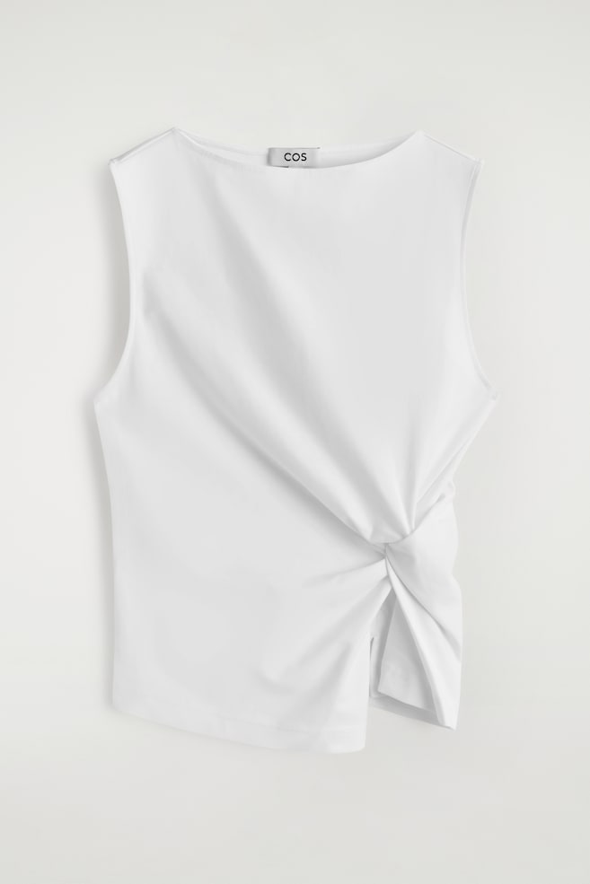 Een witte slim-fit mouwloze top toont zijn voorkant, met een boothals en een gedraaid knoopdetail aan de linkerkant van de taille, wat een geplooid, sculpturaal effect en een asymmetrische zoomlijn creëert. - 1