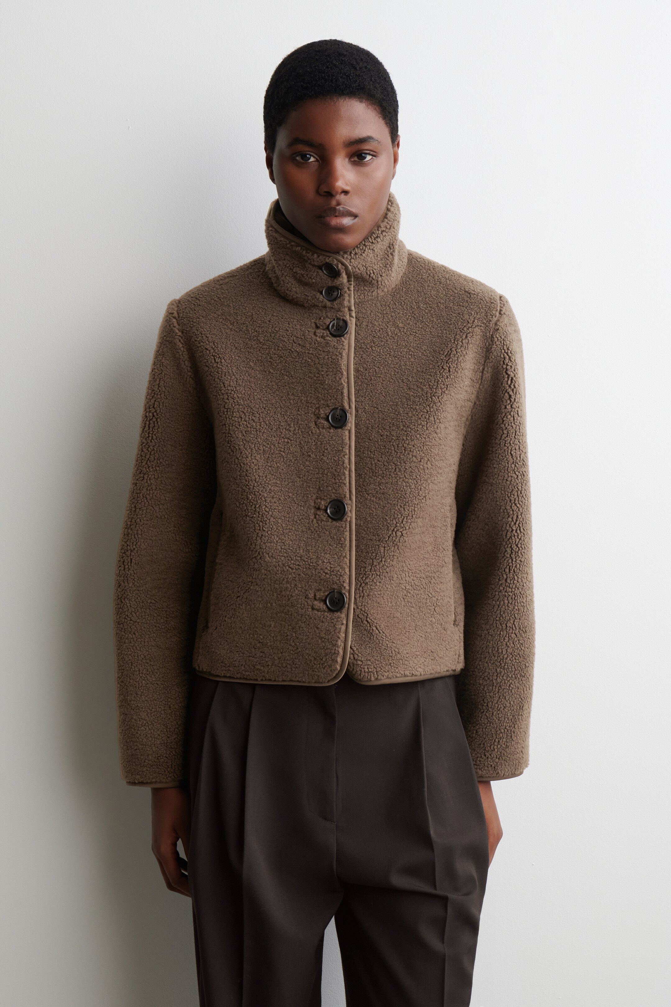 Agrandir l'image: Une veste en polaire teddy marron coupée est présentée de face. Elle présente un col montant, une fermeture frontale à quatre boutons sombres, et une bordure lisse contrastante le long de la parementure, du col et de l'ourlet.
