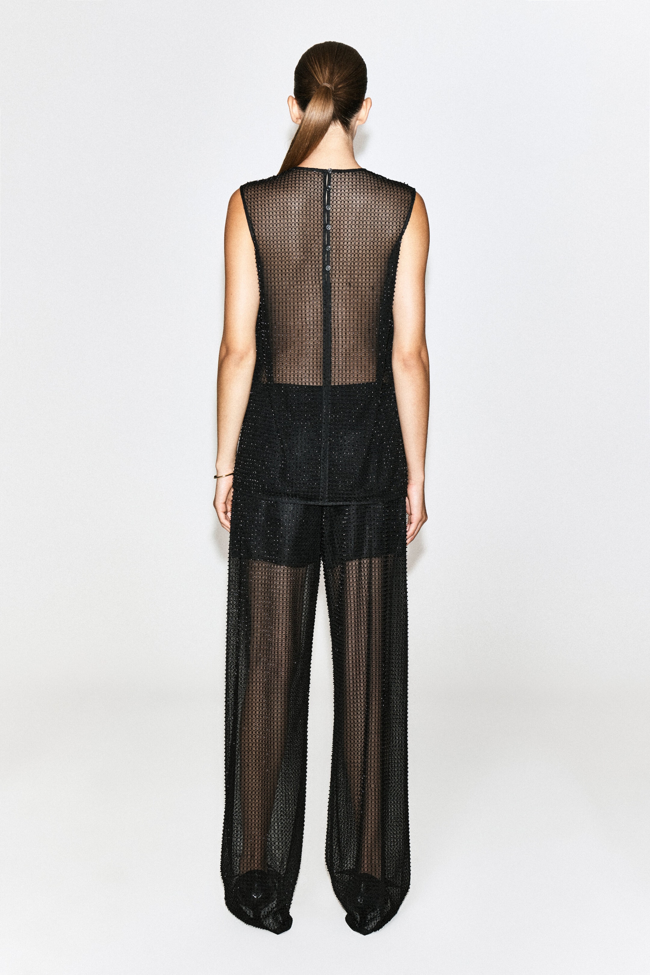 Beaded knitted trousers in a sheer, black, wide-leg style are being worn by the model in a rear view. The trousers feature an open-knit texture embellished with subtle beads, layered over solid black shorts and paired with a matching sleeveless top.
