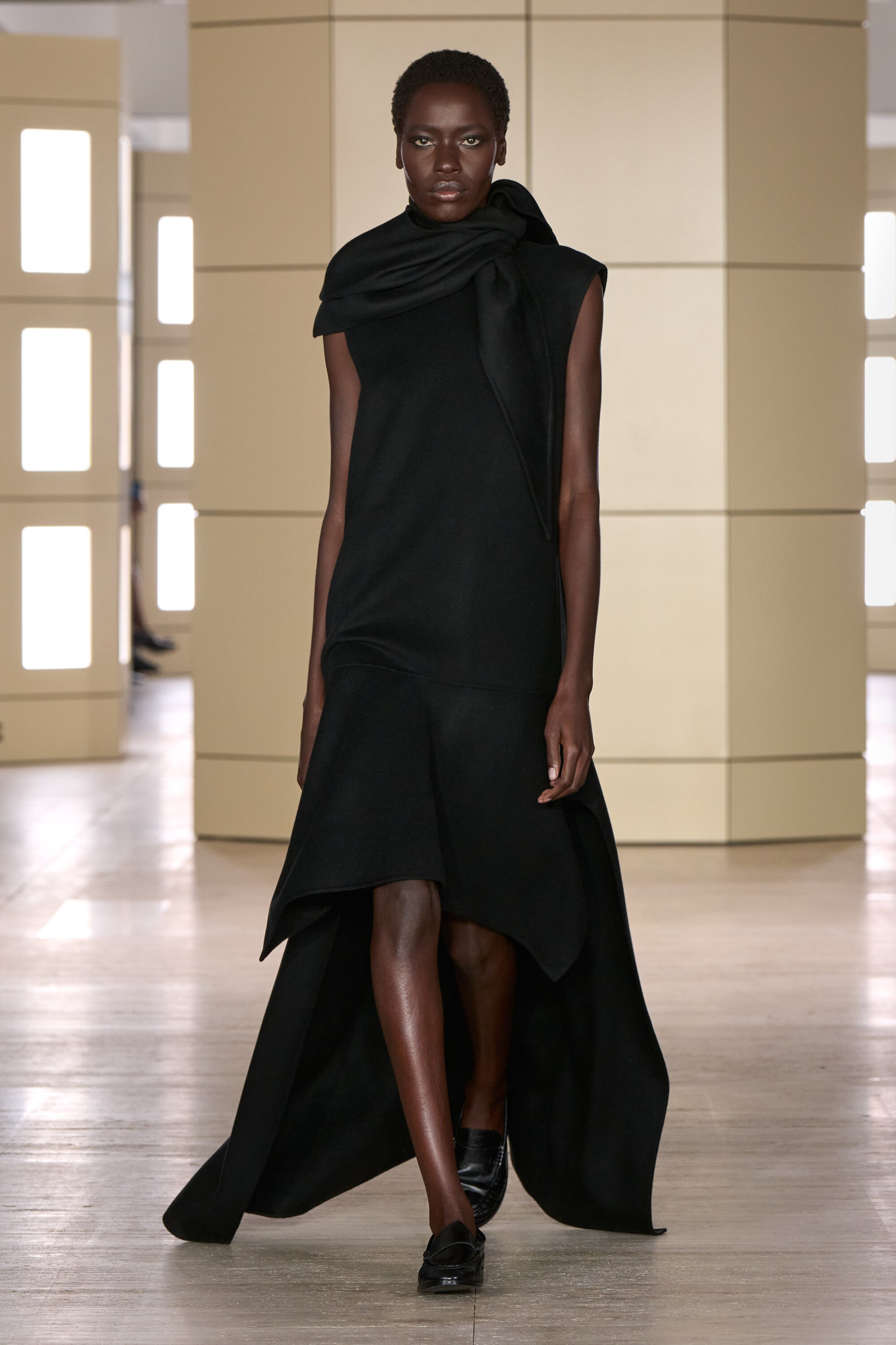 This front view showcases a black dropped-waist double-faced wool dress featuring a sleeveless silhouette with a high, draped scarf-like collar. The dress descends into a dropped waistline and flows into an asymmetric high-low hemline, draping longer at the sides and back, styled with black leather loafers. This front view showcases a black dropped-waist double-faced wool dress featuring a sleeveless silhouette with a high, draped scarf-like collar. The dress descends into a dropped waistline and flows into an asymmetric high-low hemline, draping longer at the sides and back, styled with black leather loafers.