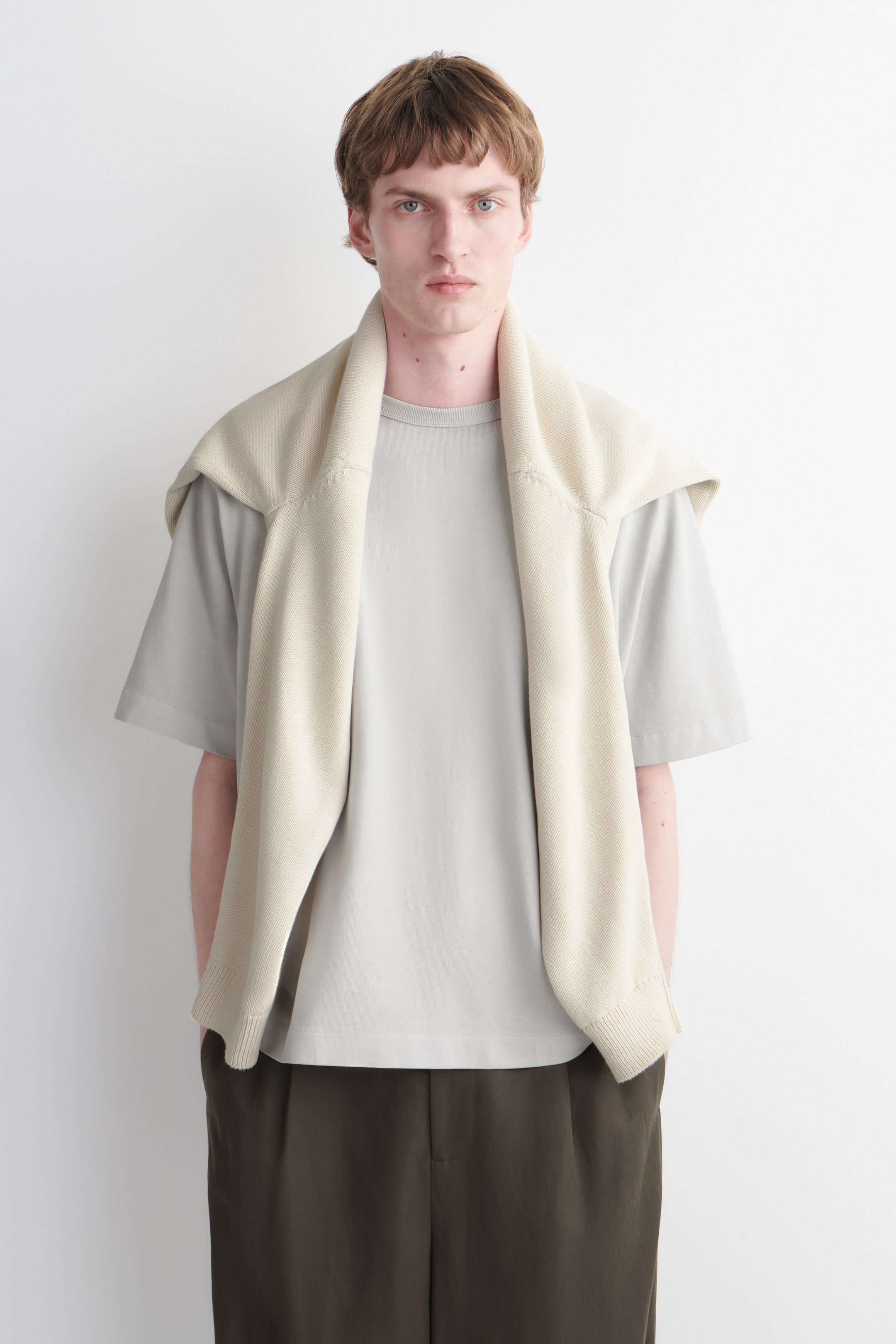 Grösseres Bild ansehen: Ein hellgraues T-Shirt mit kurzen Ärmeln und Rundhalsausschnitt wird getragen, darüber ein beige gestrickter Pullover mit gerippten Bündchen und einem gerippten Saum, lässig über die Schultern gelegt. Dunkelolivfarbene, plissierte Hosen vervollständigen das nach vorne gerichtete Outfit.