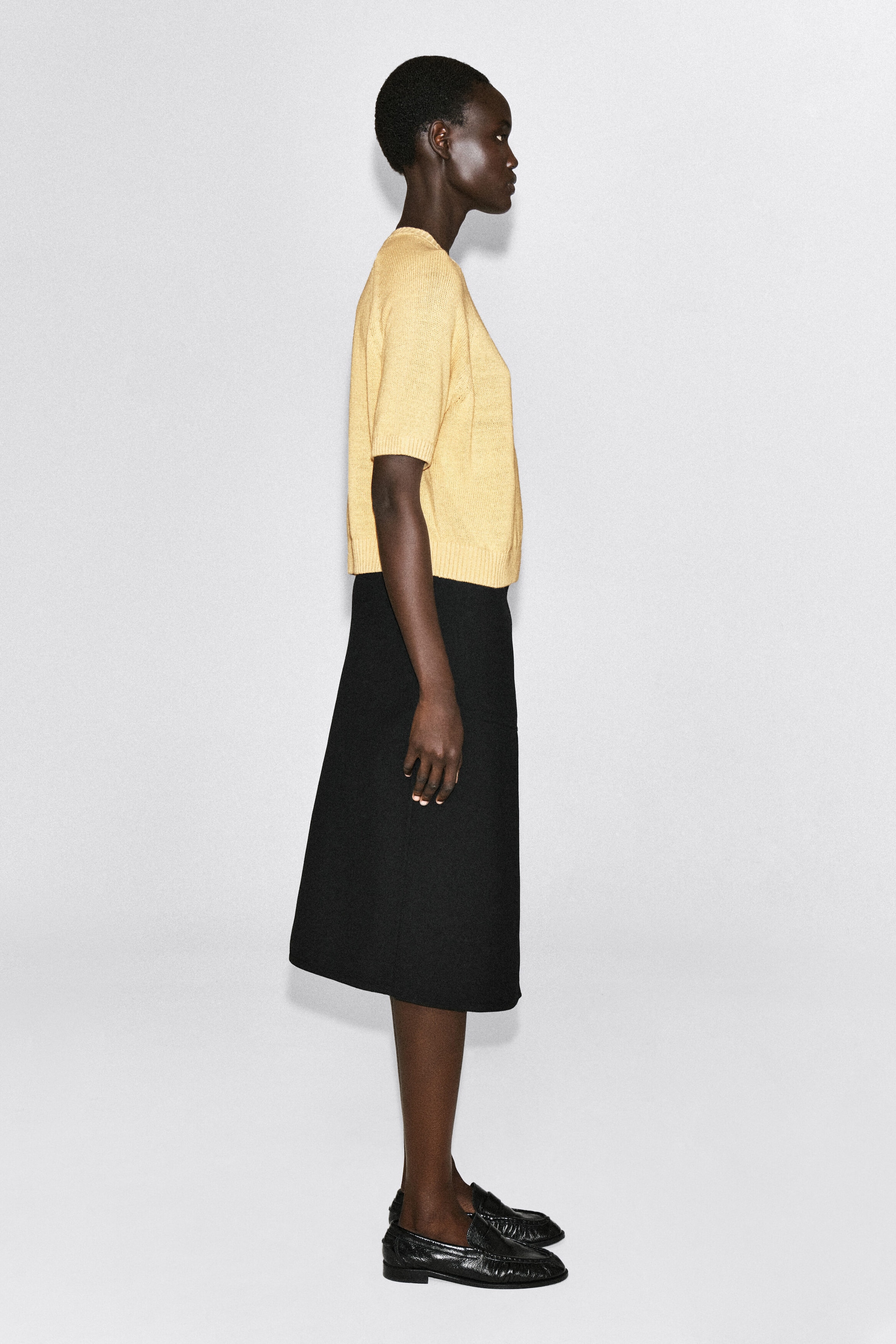 A light yellow, short-sleeved knit top featuring a crew neck, ribbed cuffs, and a ribbed hem is paired with a black knee-length A-line skirt. The model is facing right, showcasing the top’s relaxed silhouette and the skirt’s structured drape. A light yellow, short-sleeved knit top featuring a crew neck, ribbed cuffs, and a ribbed hem is paired with a black knee-length A-line skirt. The model is facing right, showcasing the top’s relaxed silhouette and the skirt’s structured drape.