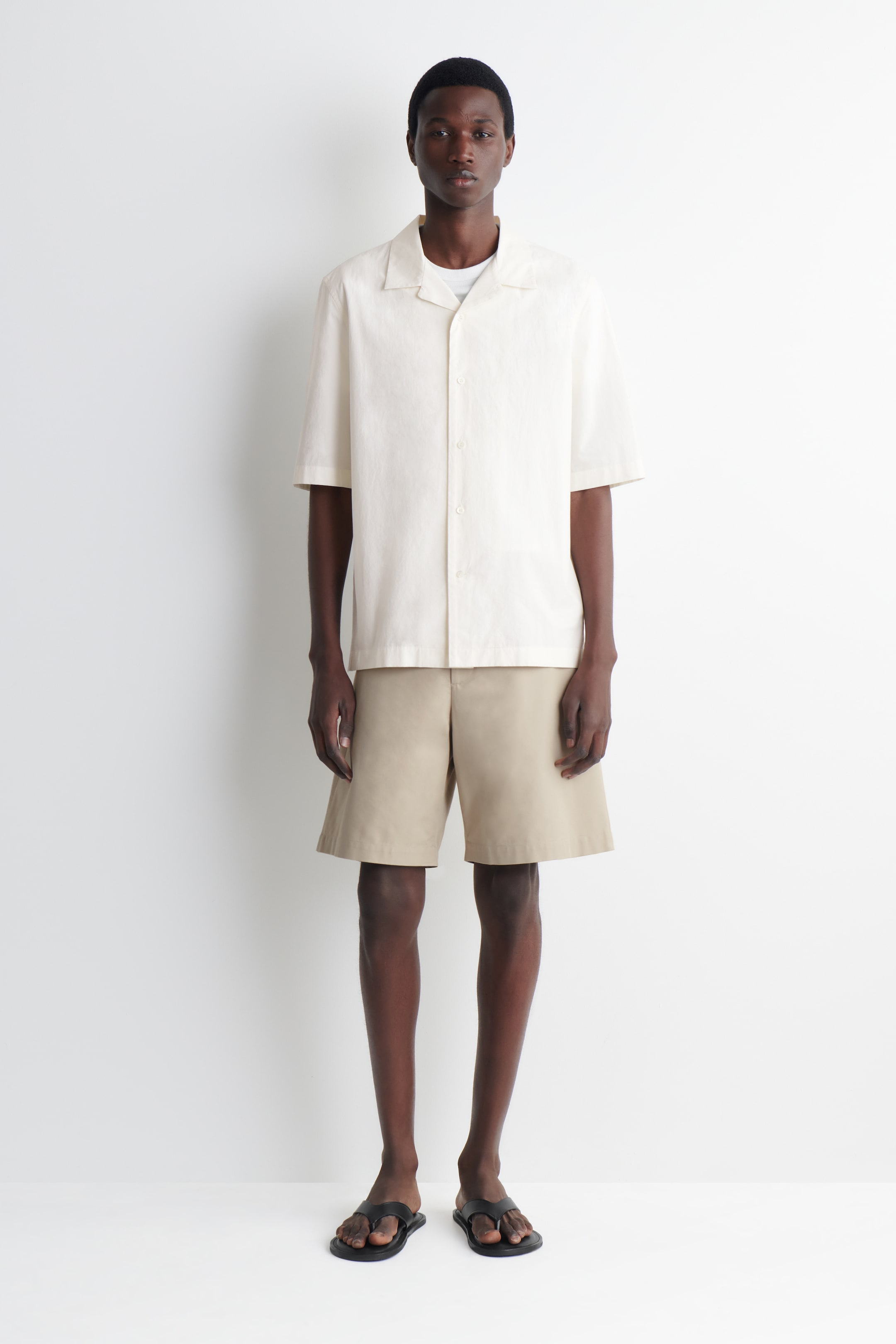 Agrandir l'image: Shorts chino beiges, avec une coupe droite et des poches latérales, portés par le mannequin. Les shorts sont assortis d'une chemise à manches courtes de couleur claire et de tongs foncées.