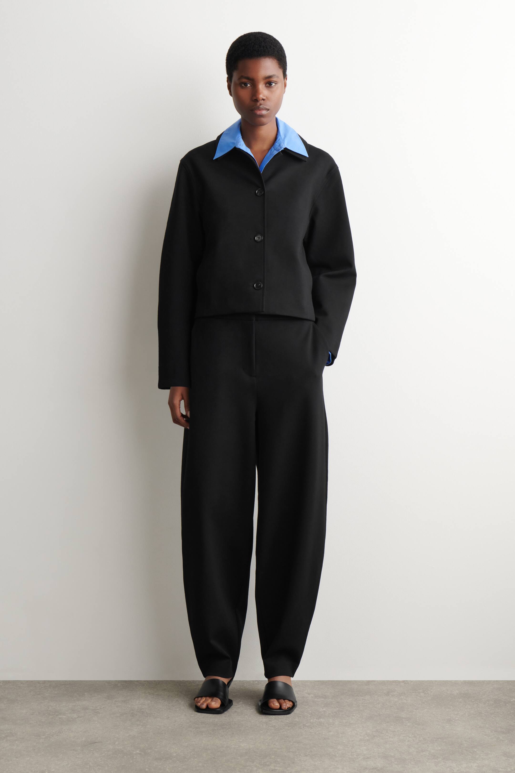 Visualizza immagine più grande: Pantaloni neri, progettati con gambe a tulipano arrotondate che si restringono alla caviglia, indossati dalla modella. La modella indossa anche una giacca corta nera sopra una camicia con colletto blu e sandali neri.