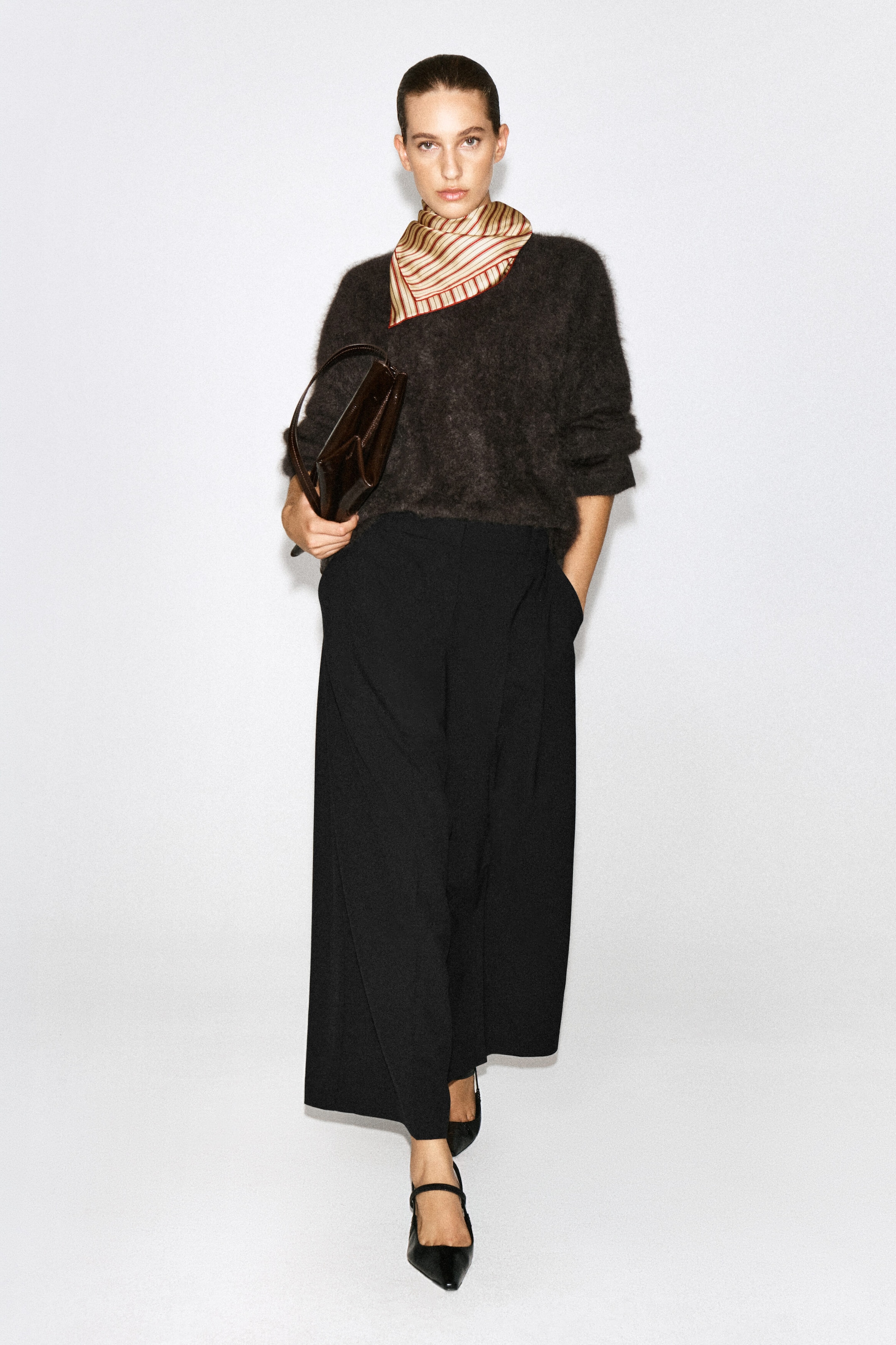 Black pleated wool-blend wide-leg trousers are being worn by the model, who is standing with one hand resting in a side pocket. The relaxed-fit, high-waisted trousers are styled with a dark fuzzy knit jumper, a striped neck scarf, and black pointed-toe flat shoes. Black pleated wool-blend wide-leg trousers are being worn by the model, who is standing with one hand resting in a side pocket. The relaxed-fit, high-waisted trousers are styled with a dark fuzzy knit jumper, a striped neck scarf, and black pointed-toe flat shoes.