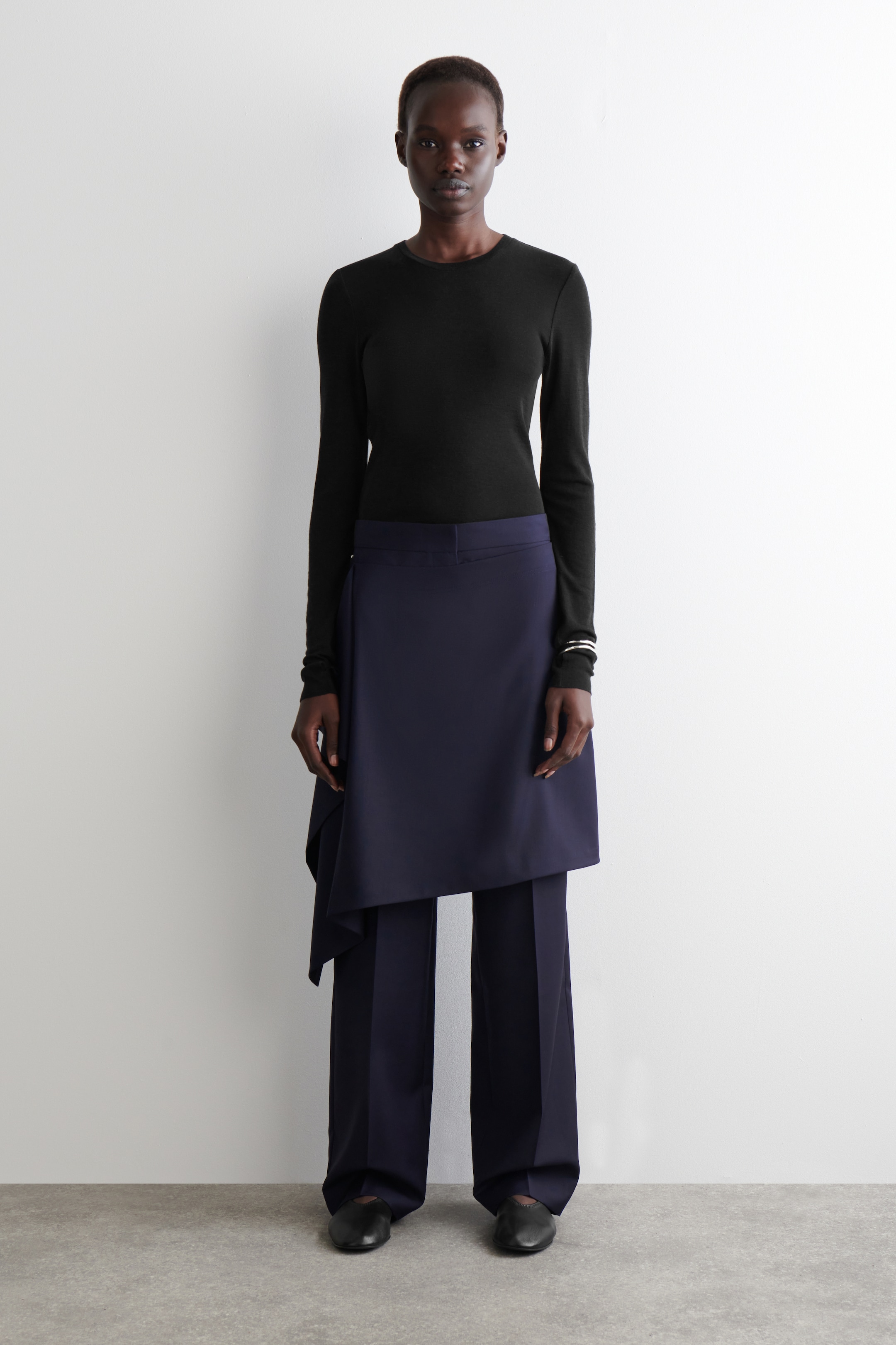 View larger image: A black long-sleeved crew-neck top is being worn, tucked into navy blue wide-leg trousers. A navy blue asymmetric wrap skirt is being layered over the trousers, with black flat shoes completing the full-front presentation.