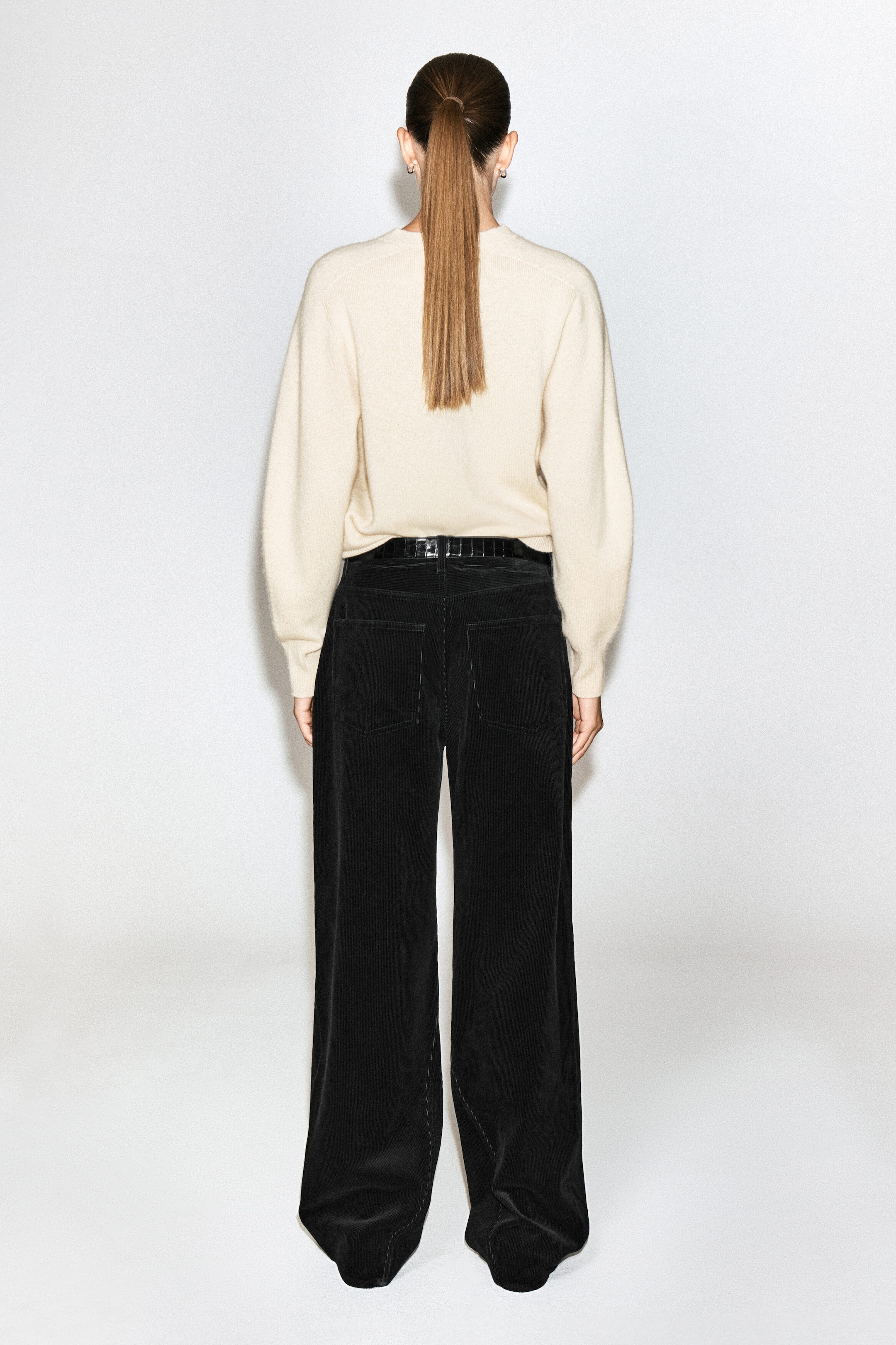 The model is wearing black corduroy straight-leg jeans, shown from the back. The high-waisted trousers feature back patch pockets and are styled with a black belt and a tucked-in cream knit jumper. The model is wearing black corduroy straight-leg jeans, shown from the back. The high-waisted trousers feature back patch pockets and are styled with a black belt and a tucked-in cream knit jumper.