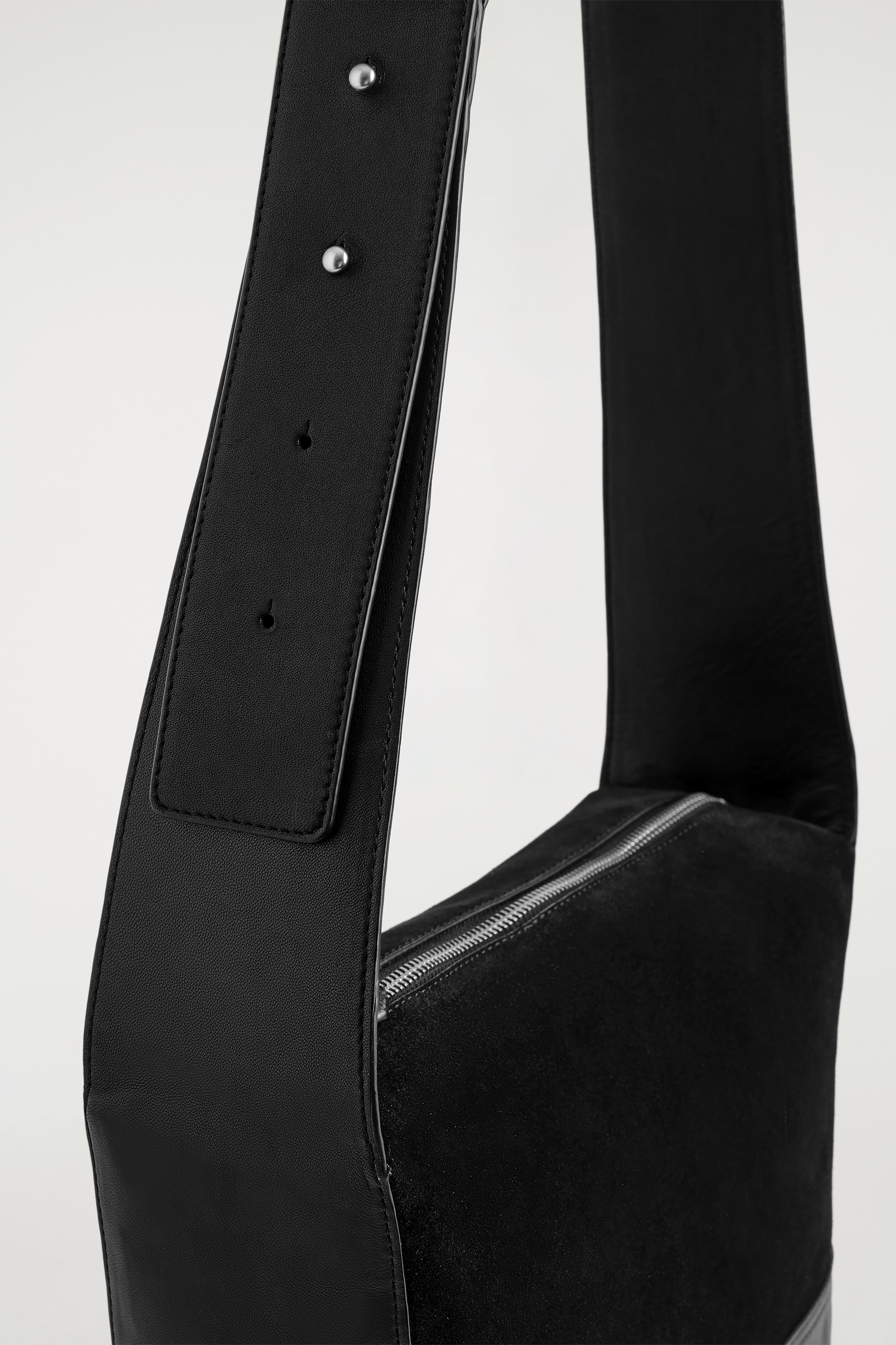Agrandir l'image: Un sac à bandoulière noir est incliné vers la droite, sa large sangle en cuir lisse présentant des coutures visibles, deux clous argentés et des trous de réglage. Le corps principal du sac est fabriqué à partir d'un matériau noir mat, peut-être semblable au daim, et est doté d'une fermeture éclair argentée qui court horizontalement sur le dessus.