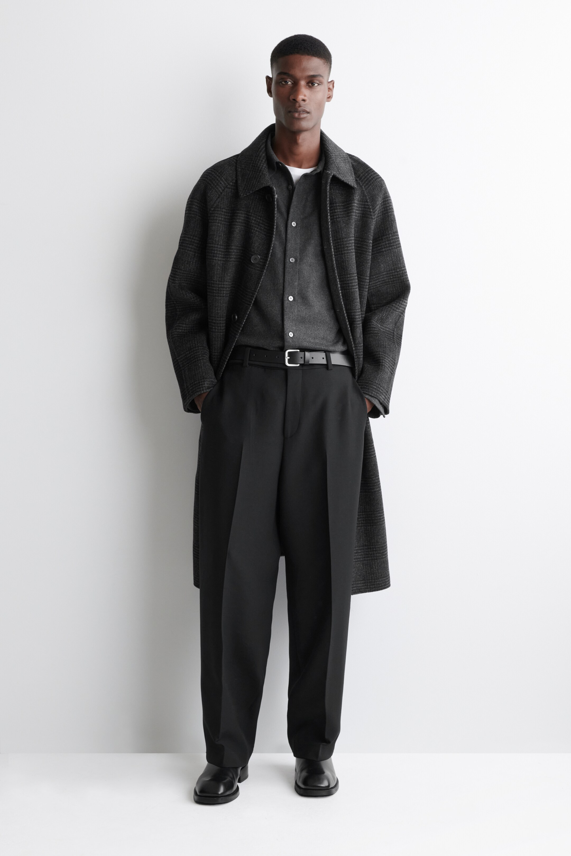 Visualizza immagine più grande: Una persona sta presentando un outfit a lunghezza intera, fronte a noi, caratterizzato da un cappotto grigio scuro a quadri con ampi revers e maniche raglan, stratificato sopra una camicia a bottoni antracite e una maglietta bianca. Indossano anche pantaloni neri dal taglio ampio con pieghe, assicurati da una cintura di pelle nera con fibbia argentata, e scarpe eleganti a punta quadrata in pelle nera.