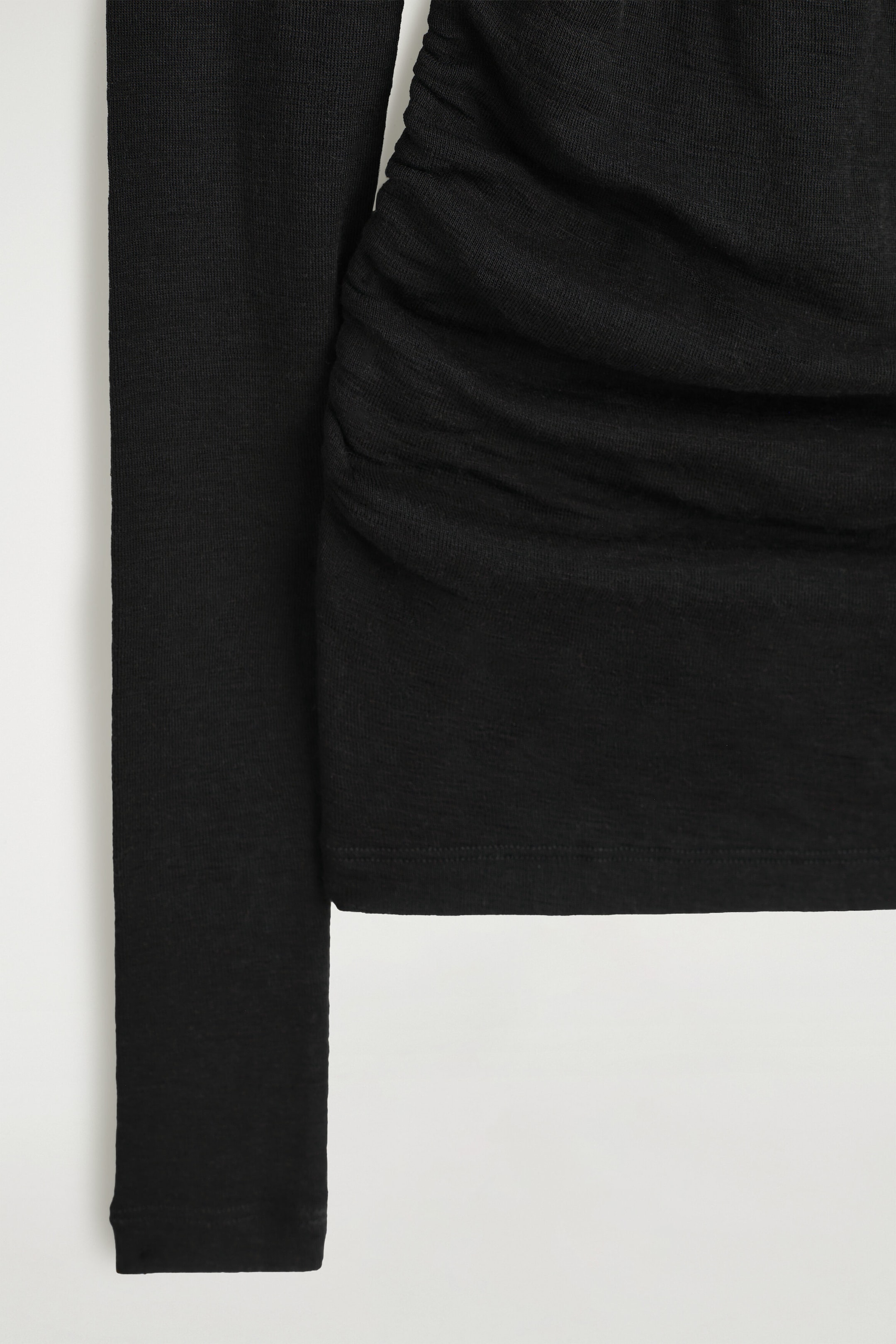 This black long-sleeved top showcases its fine-knit texture and detailed construction in a close-up view. The garment features a slim-cut sleeve alongside gathered, ruched fabric that creates draped horizontal folds across the body. This black long-sleeved top showcases its fine-knit texture and detailed construction in a close-up view. The garment features a slim-cut sleeve alongside gathered, ruched fabric that creates draped horizontal folds across the body.